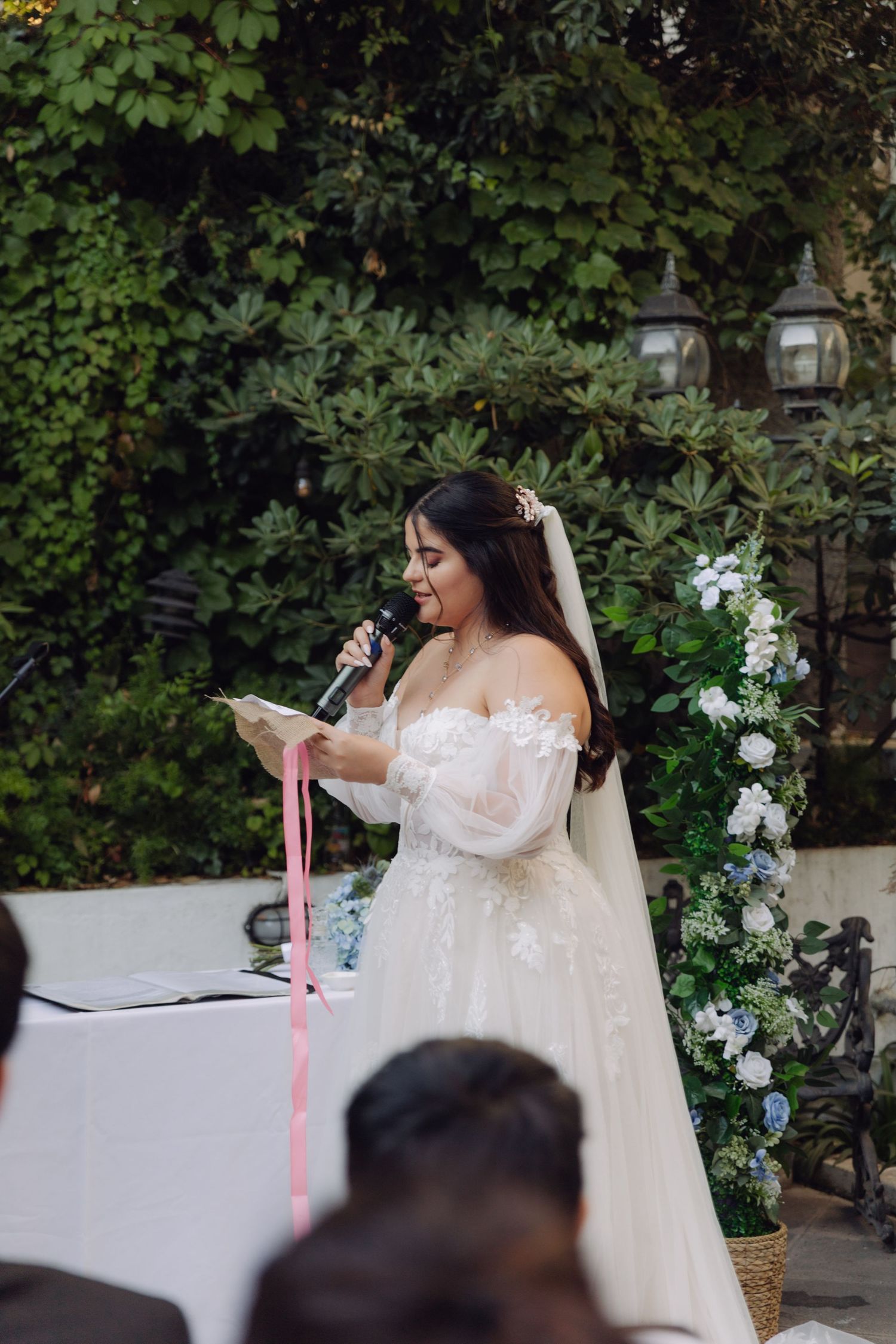 Bride in off-shoulder lace wedding gown reads vows at floral-decorated outdoor ceremony altar.