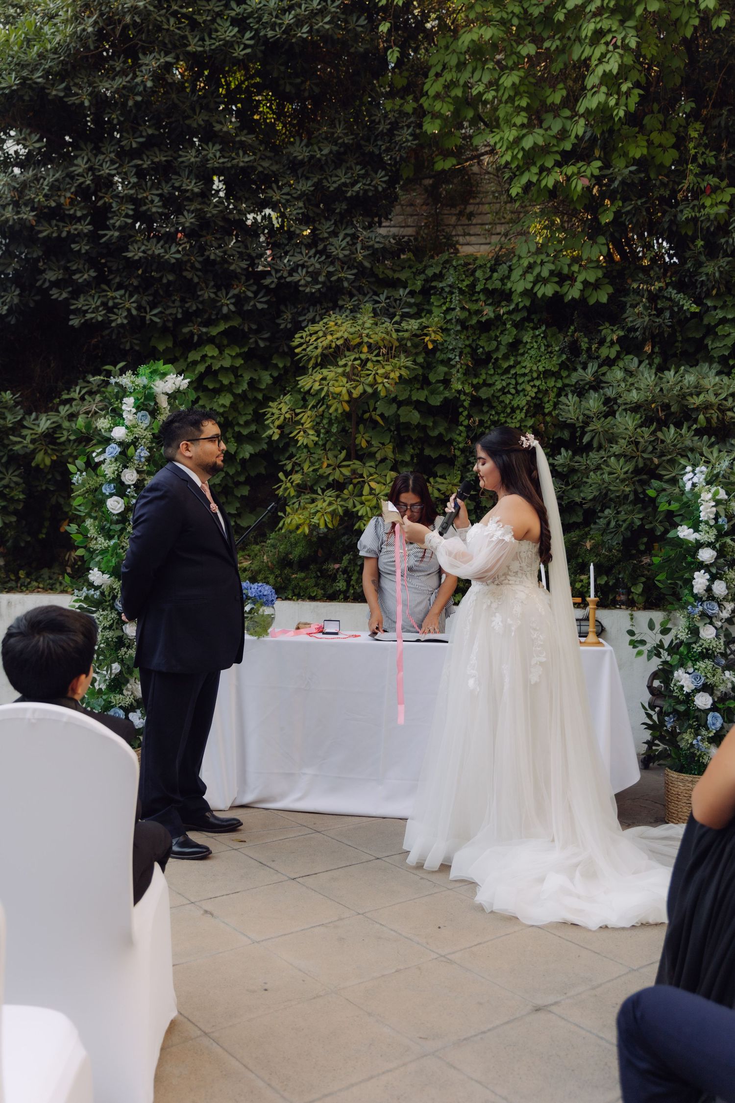Bride holds pink ribbon during outdoor wedding handfasting ceremony with groom in navy suit.