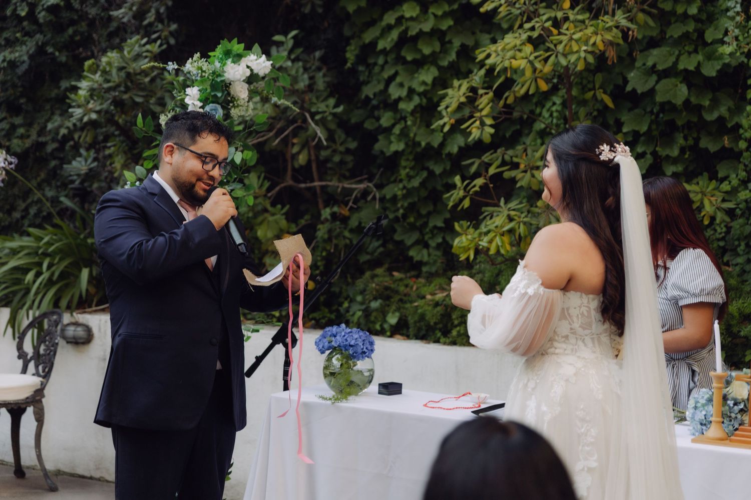Groom in navy suit holds pink ribbon during handfasting ceremony while bride stands opposite.