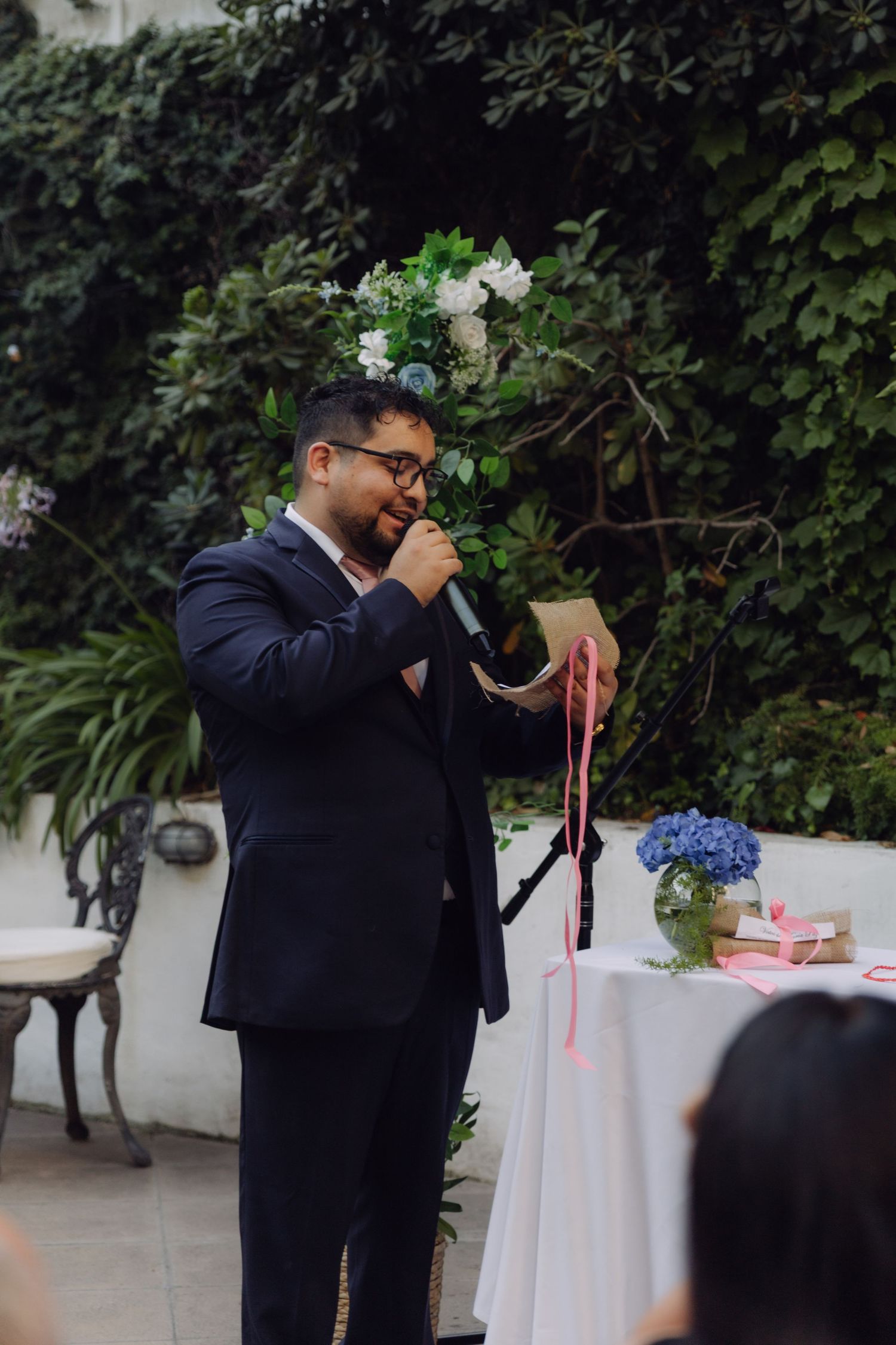 Groom in navy suit smiles while reading vows into microphone at outdoor garden wedding.