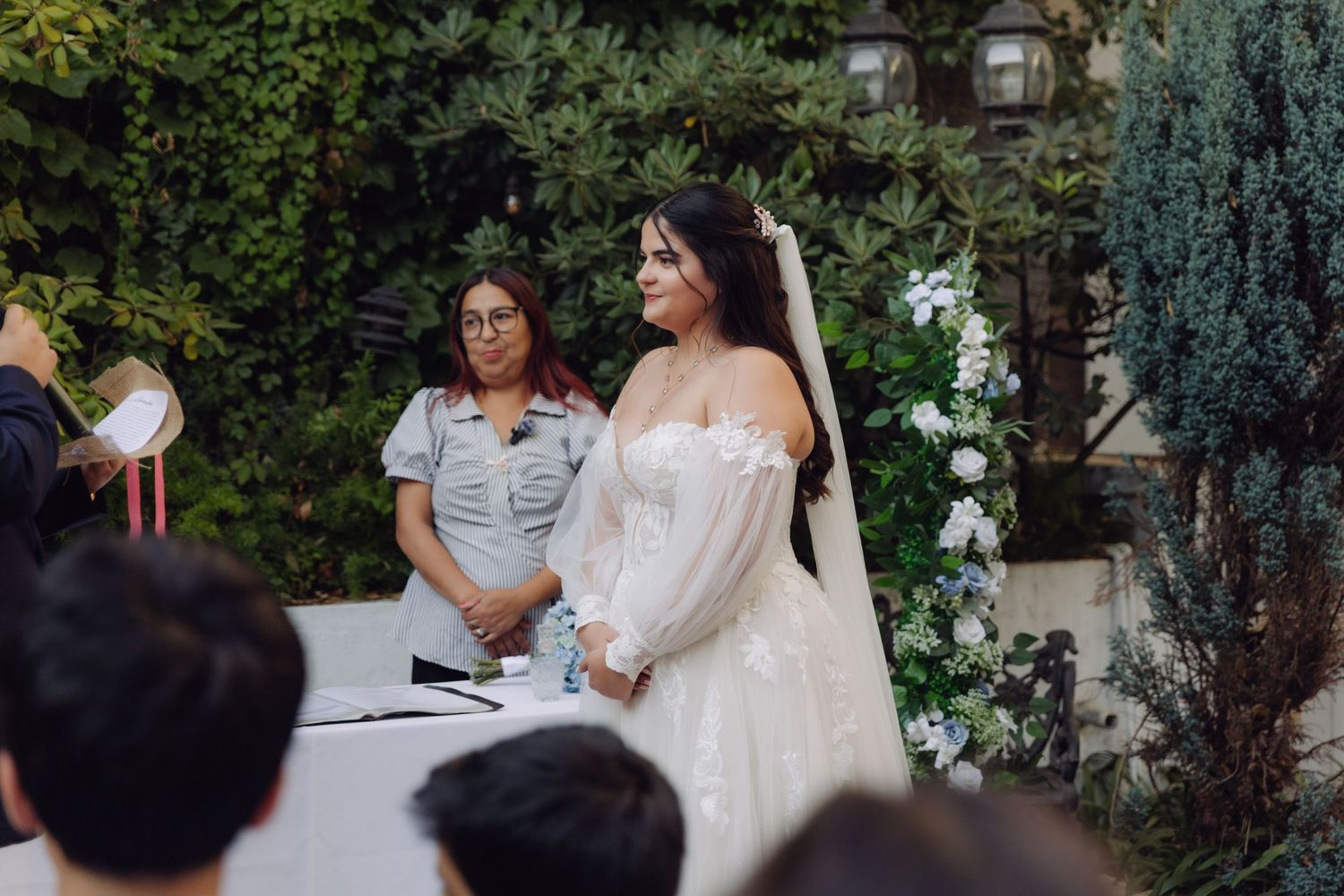 Bride in off-shoulder lace wedding gown smiling during outdoor garden ceremony.