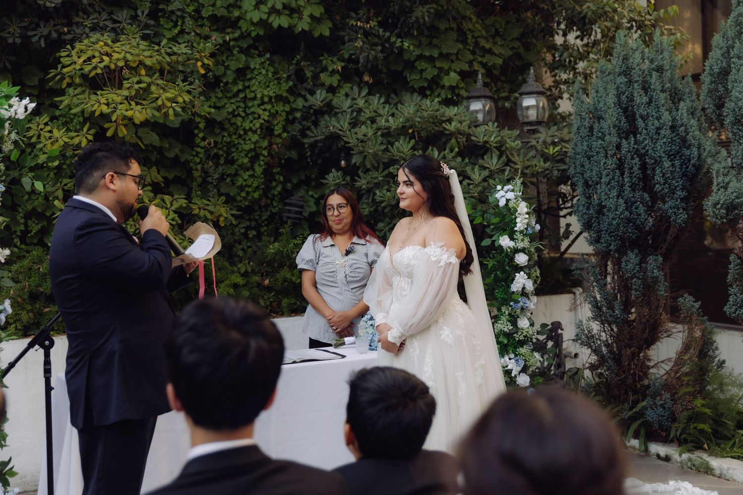 Groom in black suit reading vows to bride in white gown at outdoor wedding ceremony.
