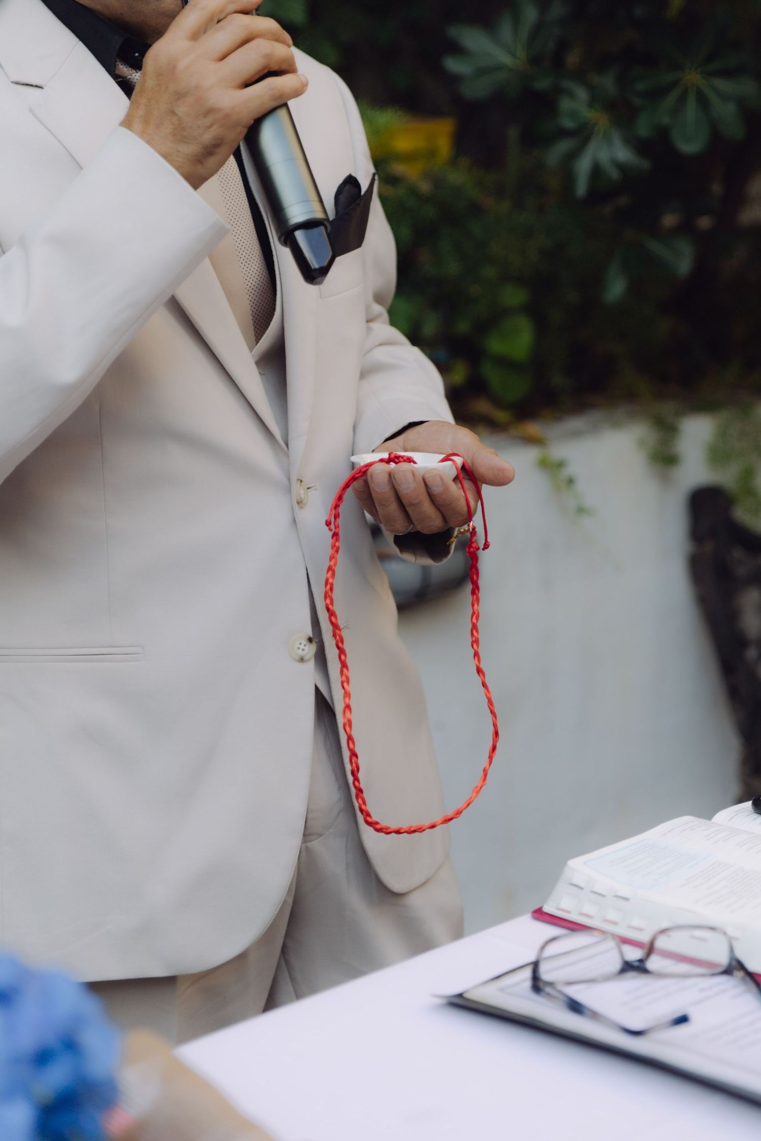 Officiant in beige suit holding red cord and microphone during wedding ceremony.