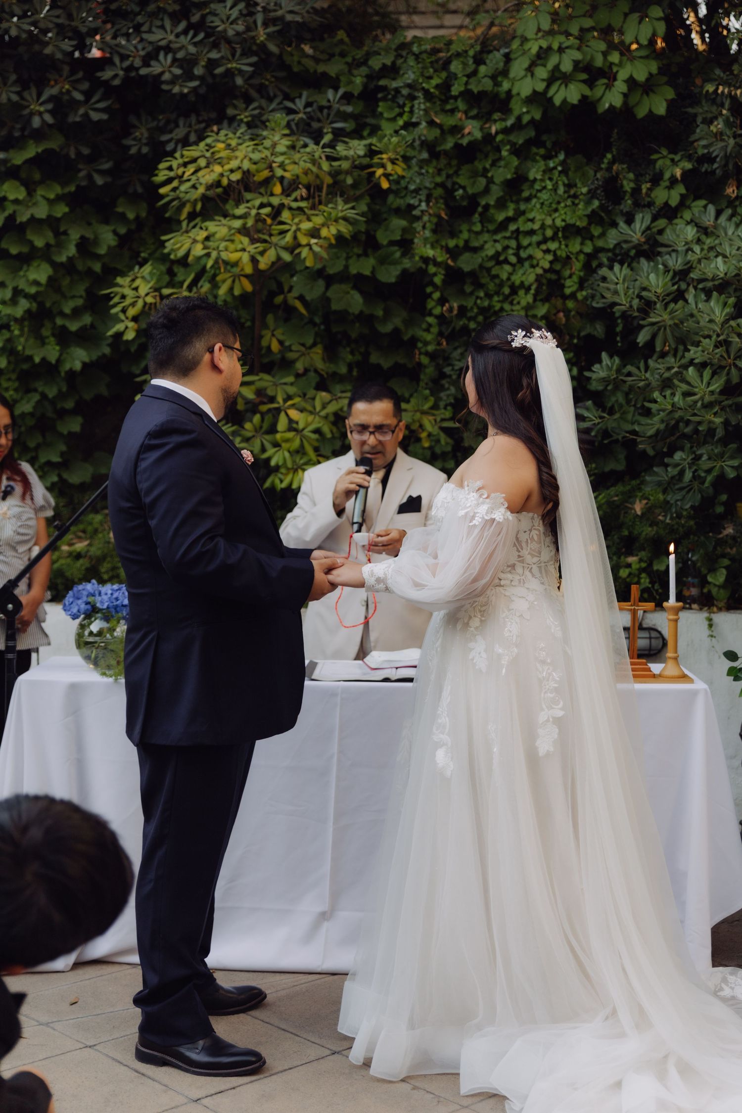 Groom placing ring on bride's finger during outdoor garden wedding ceremony with officiant.