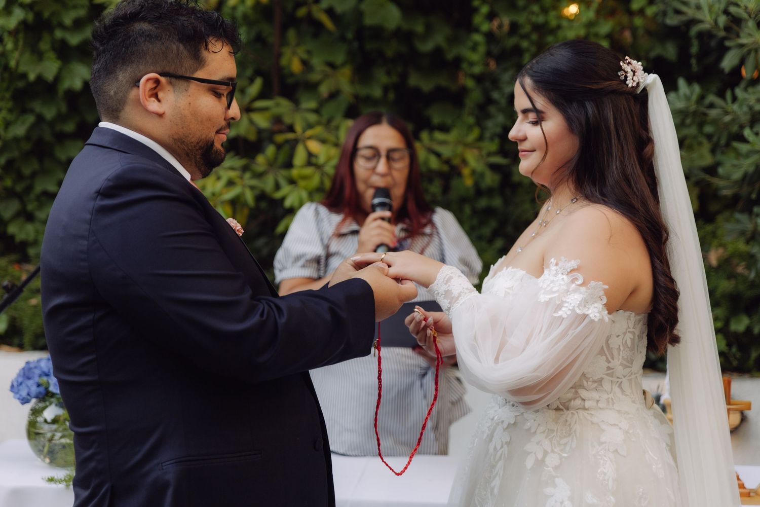Groom placing wedding ring on smiling bride's finger during outdoor garden ceremony.