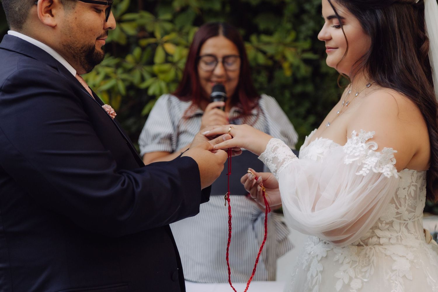 Groom sliding wedding ring onto bride's finger with red cord during outdoor ceremony.