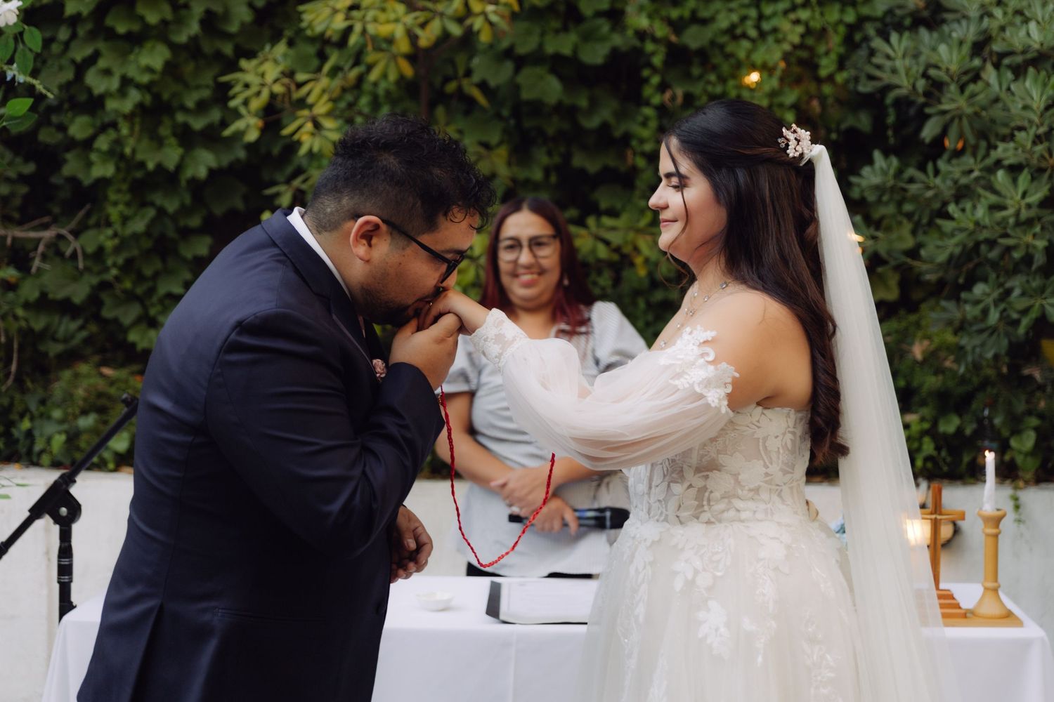 Groom kissing bride's hand during outdoor garden wedding ceremony with officiant behind them.