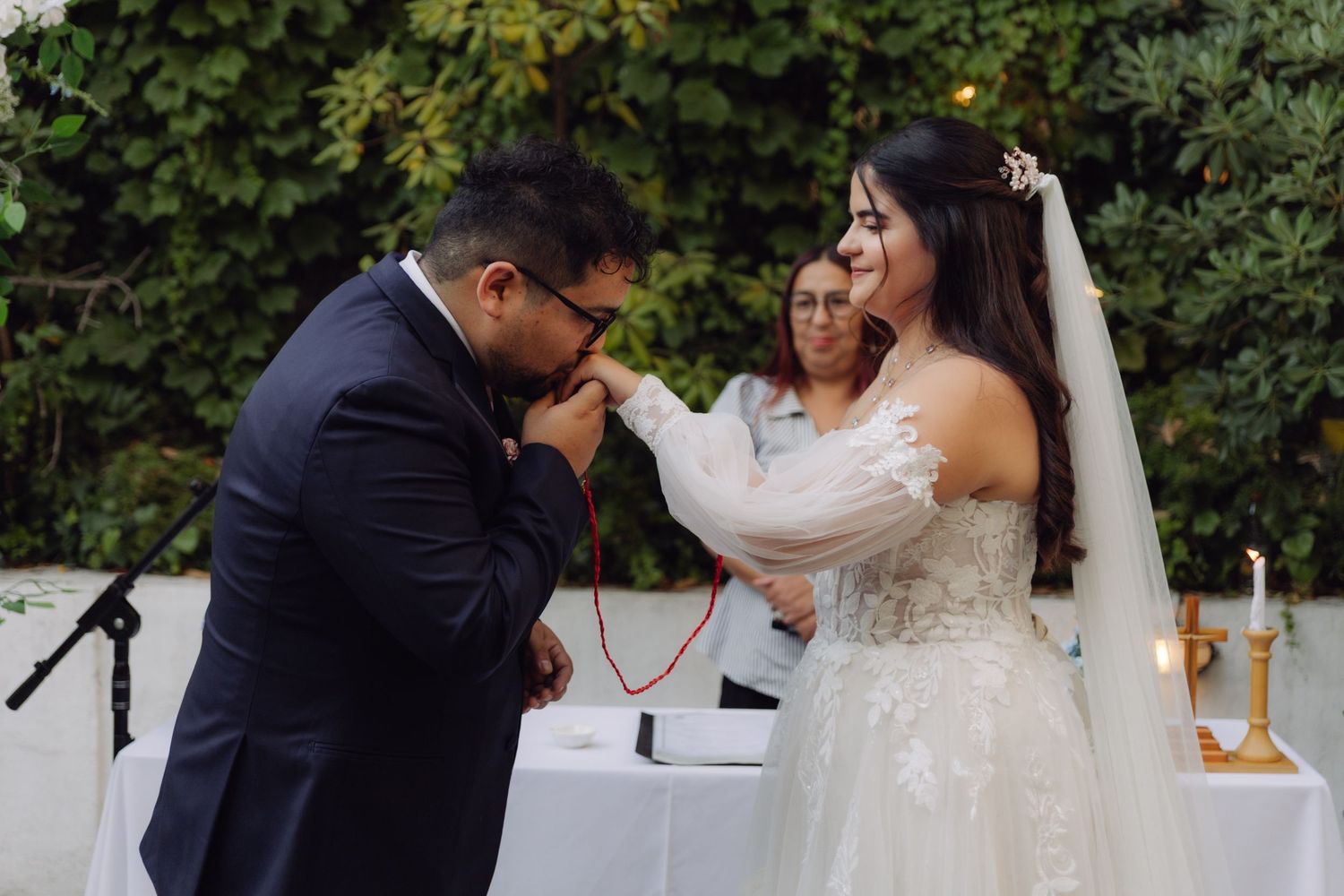 Groom bowing to kiss bride's hand during romantic outdoor garden wedding ceremony.