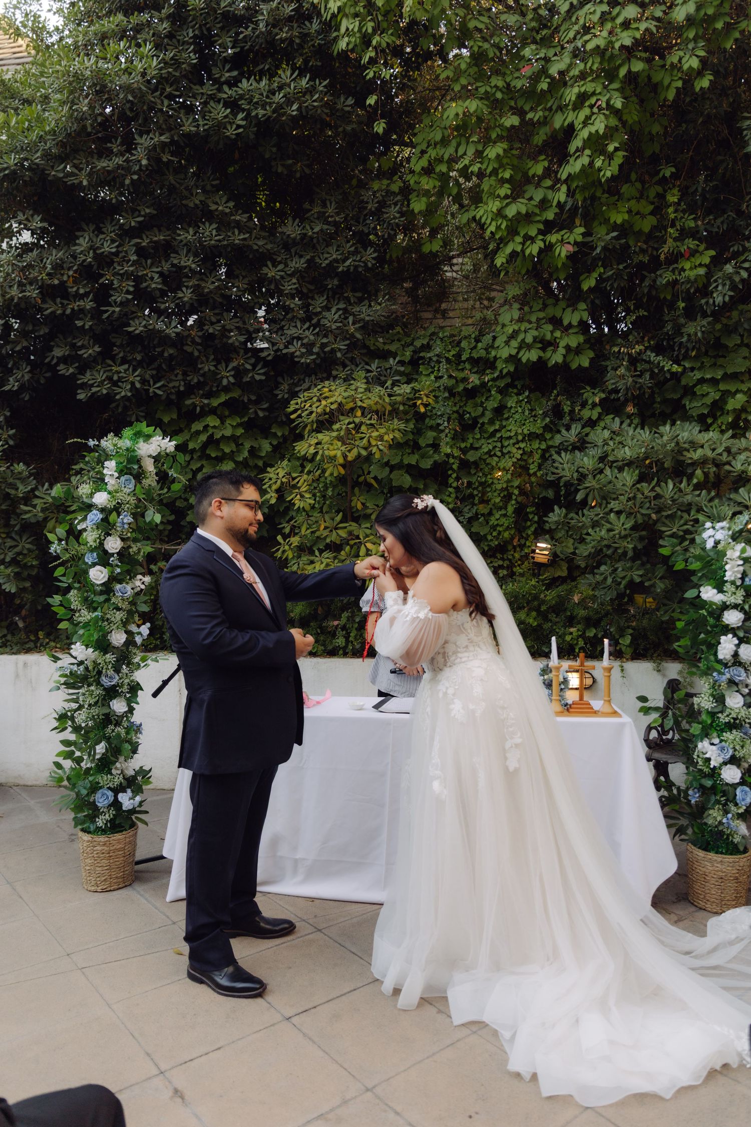 Groom in navy suit places ring on bride's finger during outdoor wedding ceremony with floral arch backdrop.