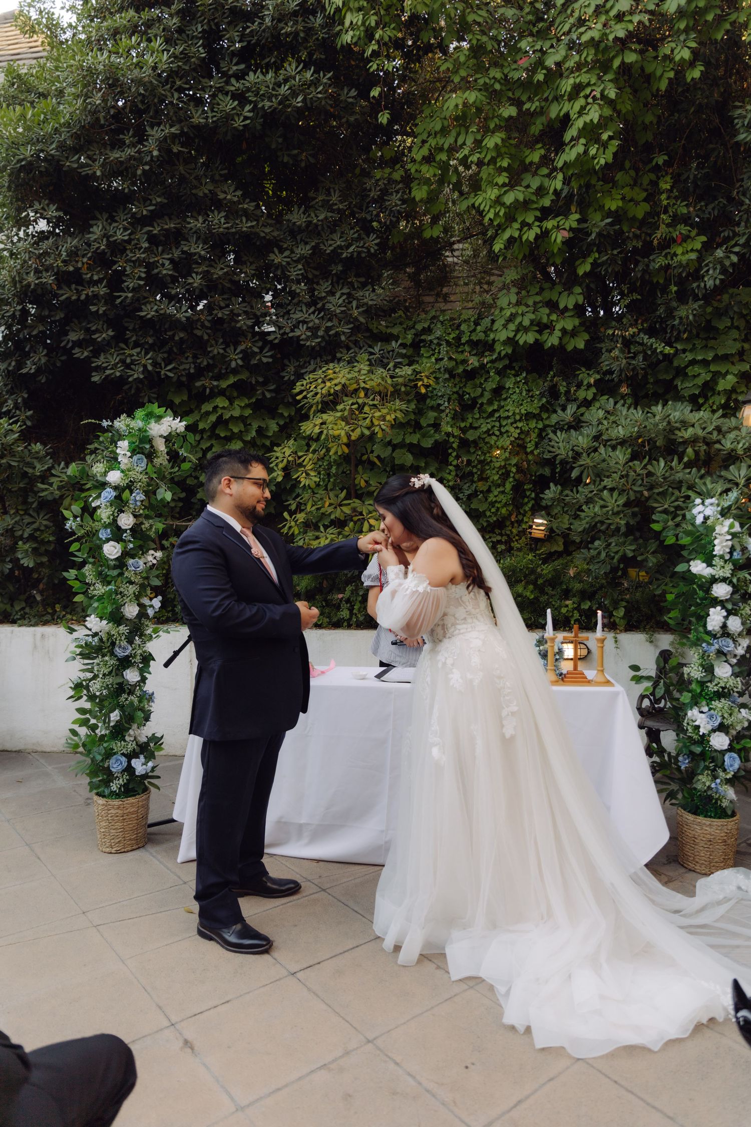 Bride in white gown and veil exchanges rings with groom during elegant outdoor garden wedding ceremony.