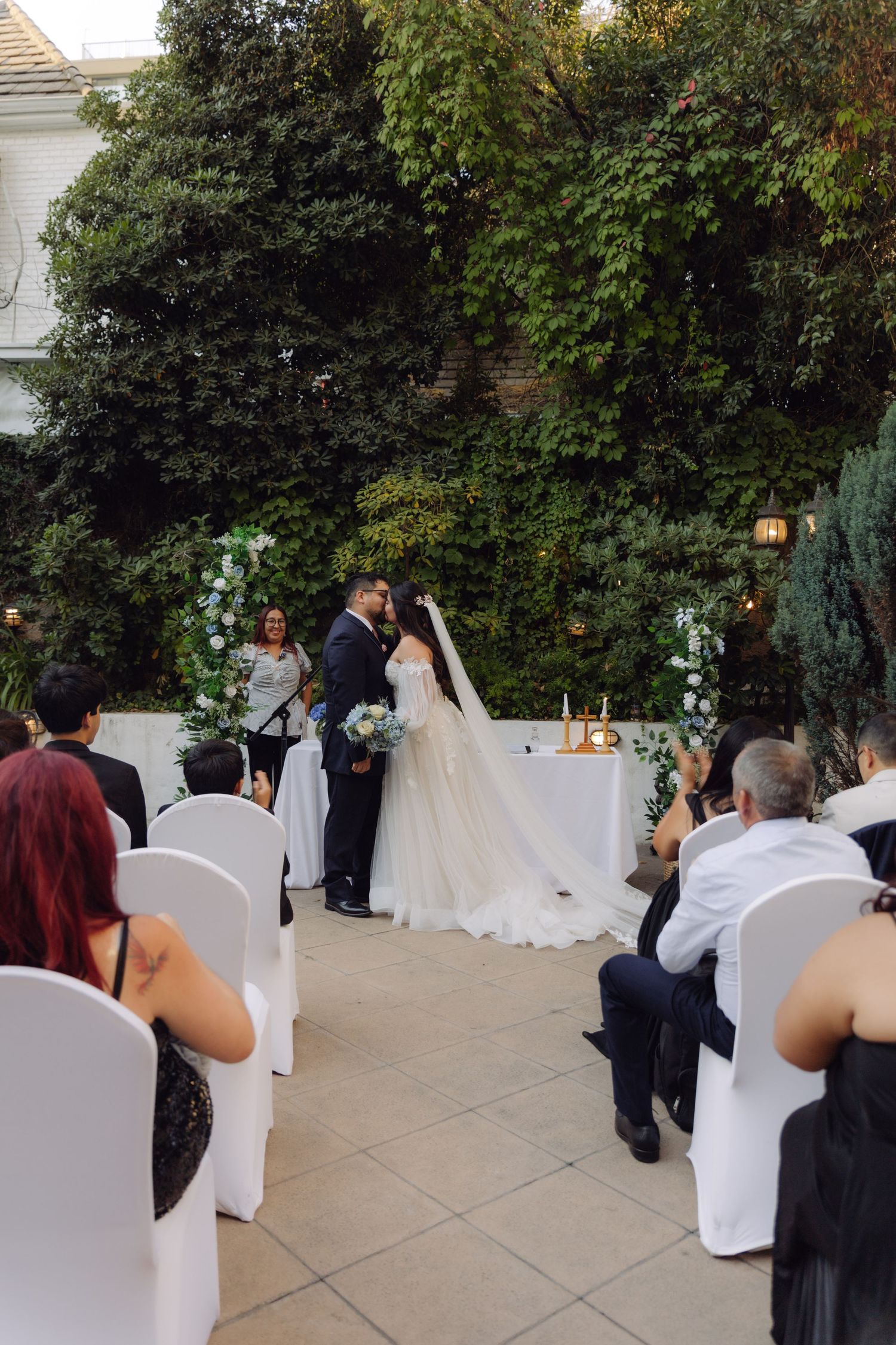 Newlyweds share first kiss at outdoor wedding ceremony as guests applaud in white chair seating.