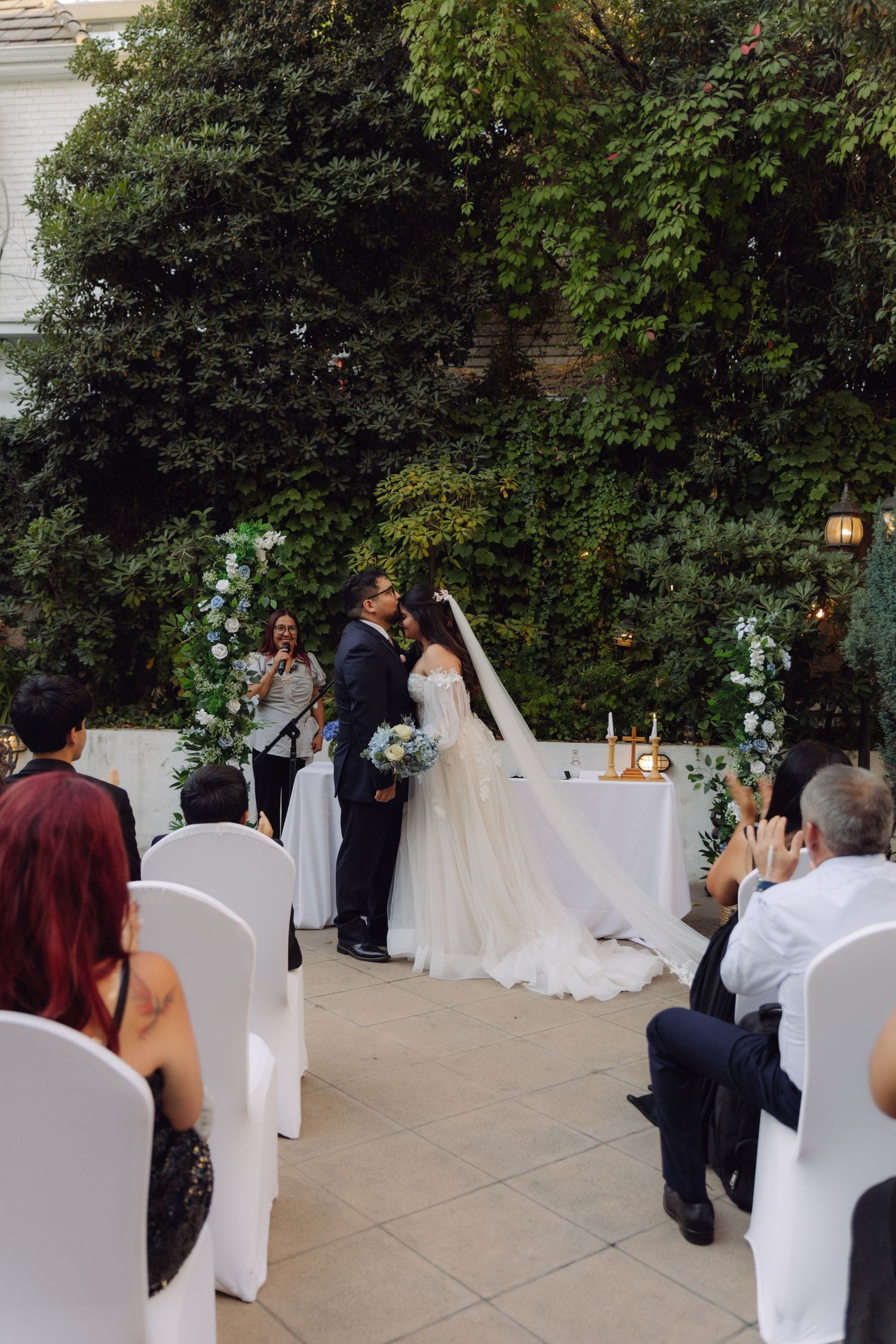 Bride and groom kiss at altar during outdoor garden wedding ceremony surrounded by seated guests.