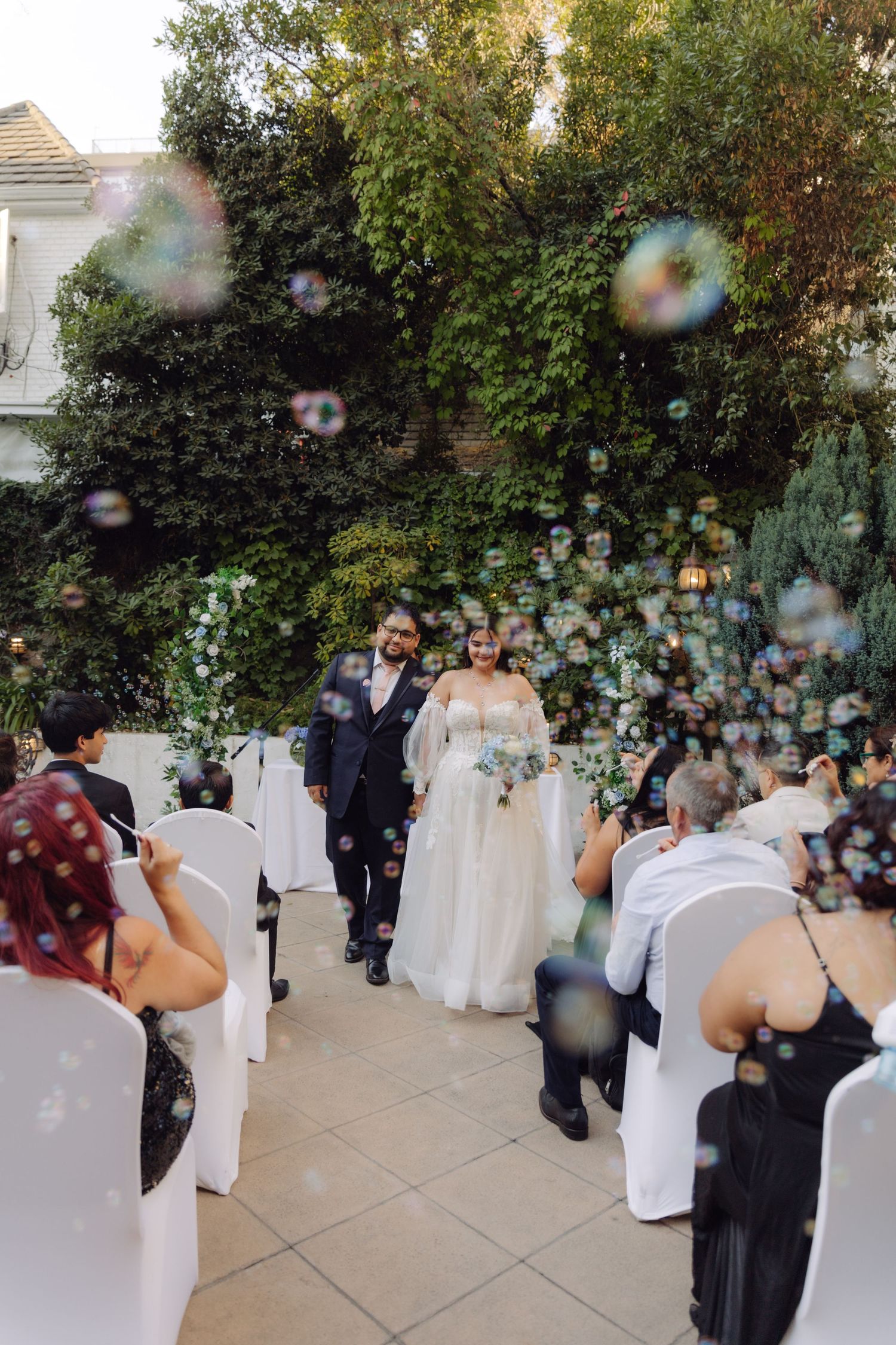 Newlyweds walk down aisle through romantic soap bubbles after outdoor garden wedding ceremony.