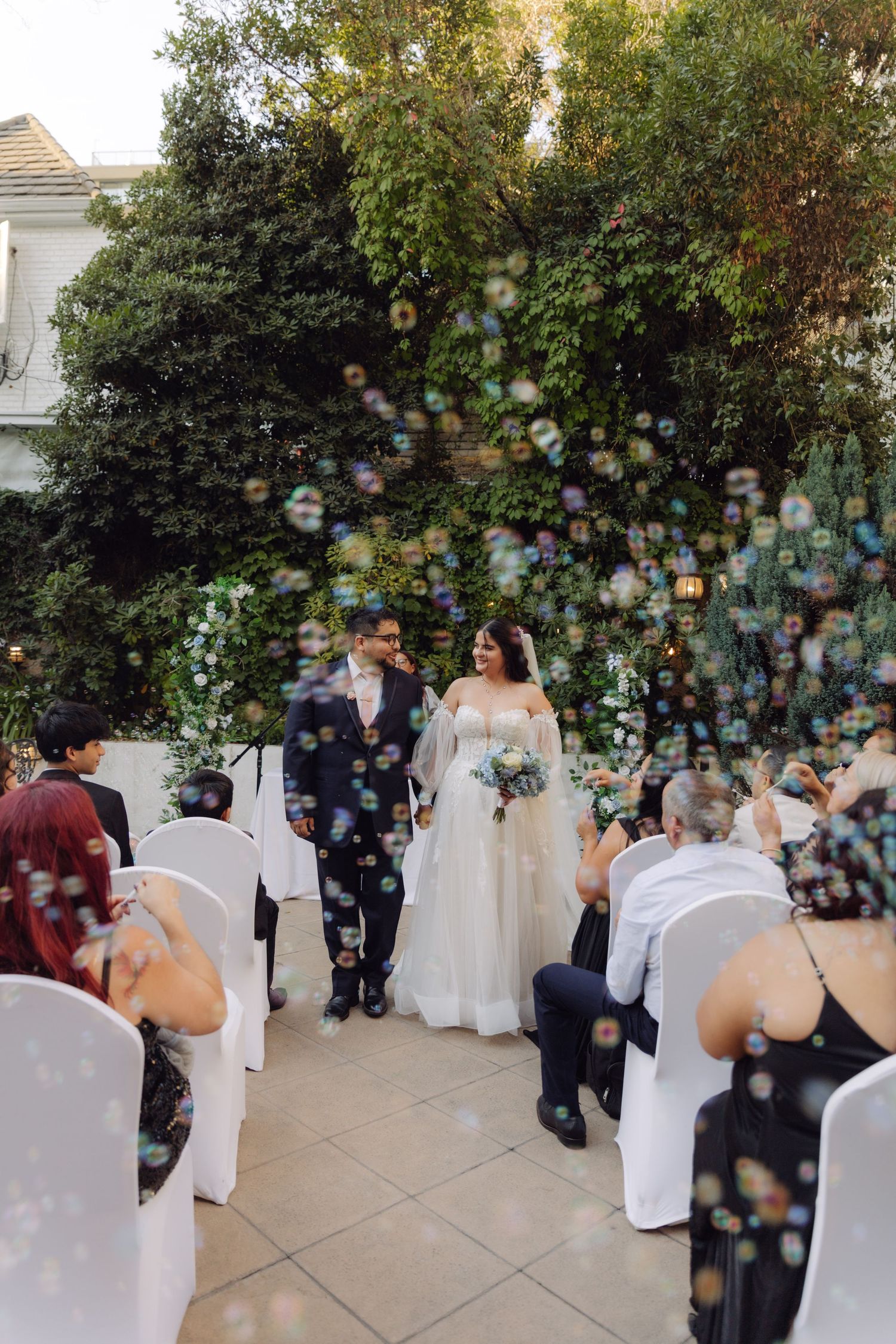 Bride and groom walking down the aisle surrounded by floating bubbles at an outdoor garden wedding.