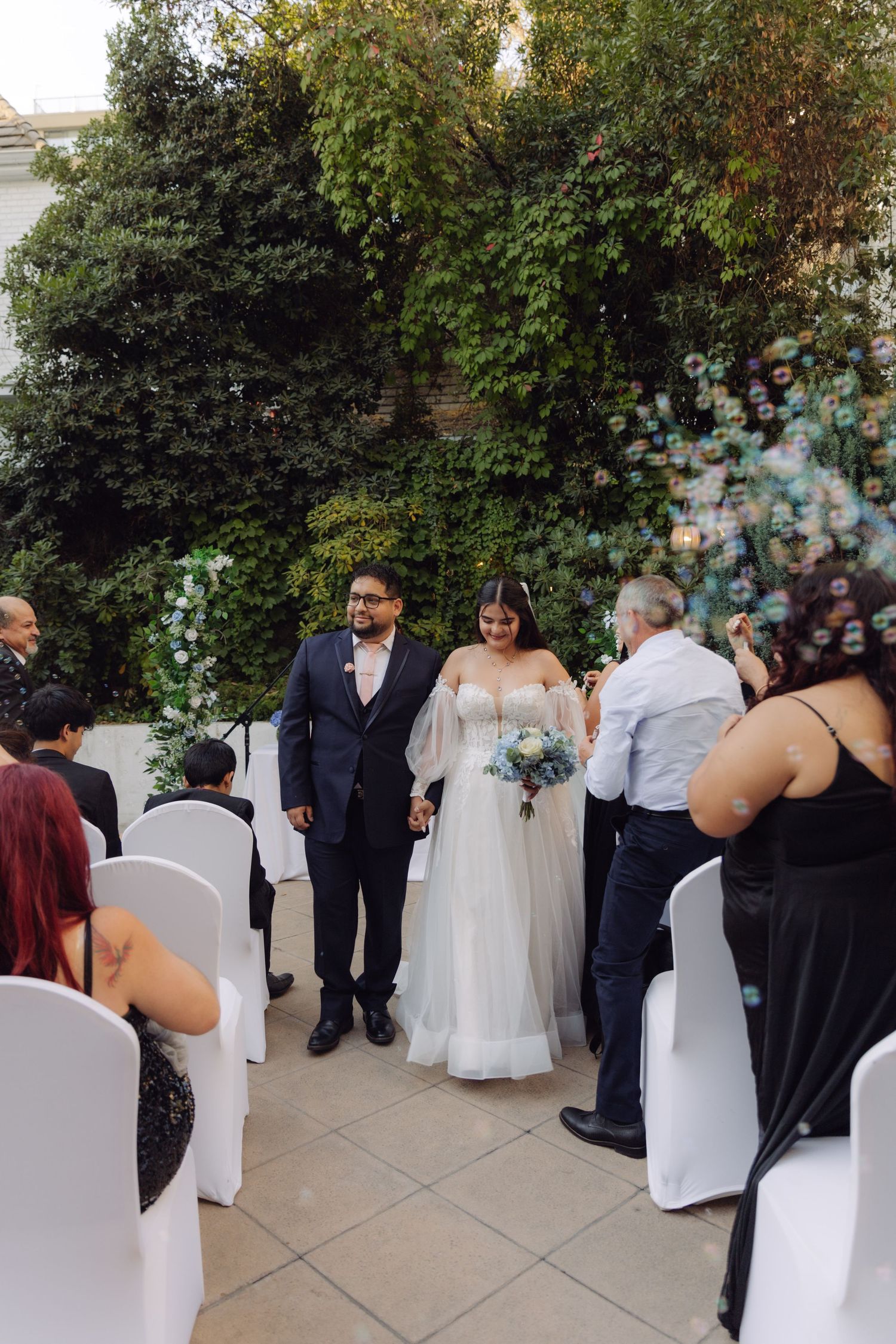 Bride and groom holding hands while walking down the aisle at an elegant outdoor garden ceremony.