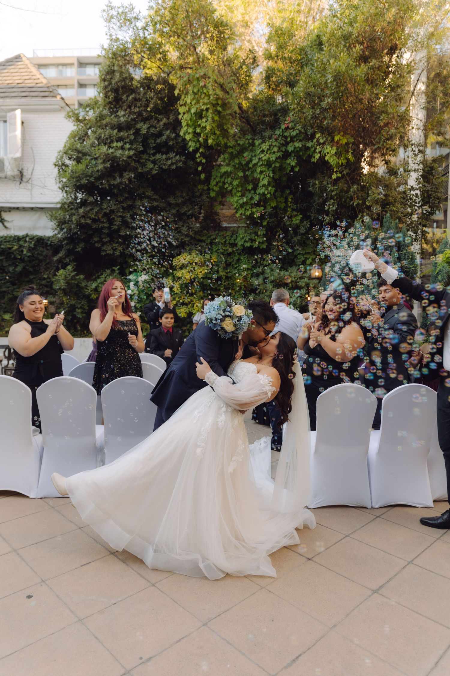 Bride and groom sharing a romantic dip kiss surrounded by cheering guests and bubbles at outdoor wedding.