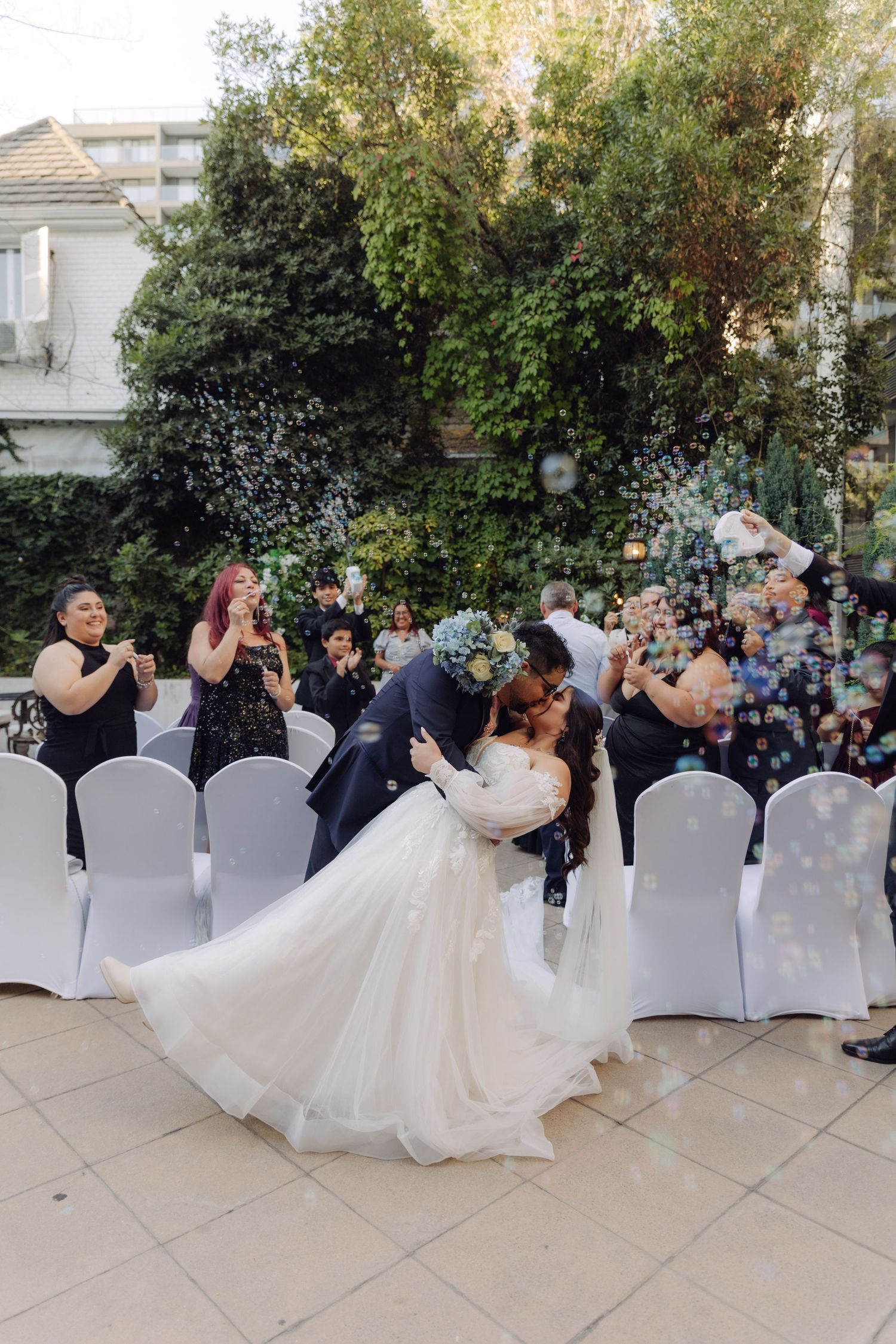 Groom dipping bride in a joyful kiss as guests celebrate with bubbles at an outdoor garden wedding.