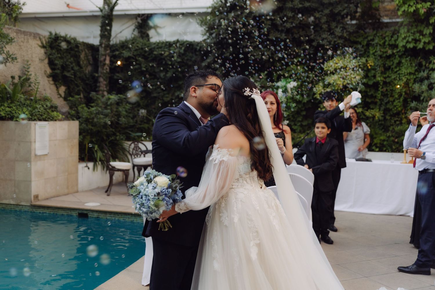 Bride and groom kissing near a pool surrounded by bubbles and guests at an outdoor wedding reception.