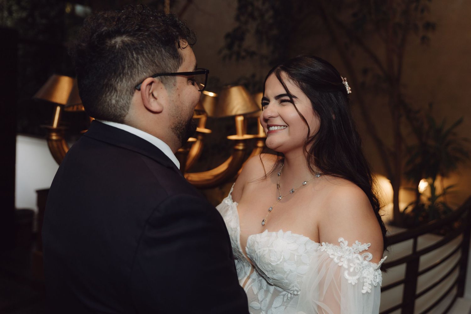 Smiling bride in off-shoulder lace gown gazes at groom during romantic wedding reception.