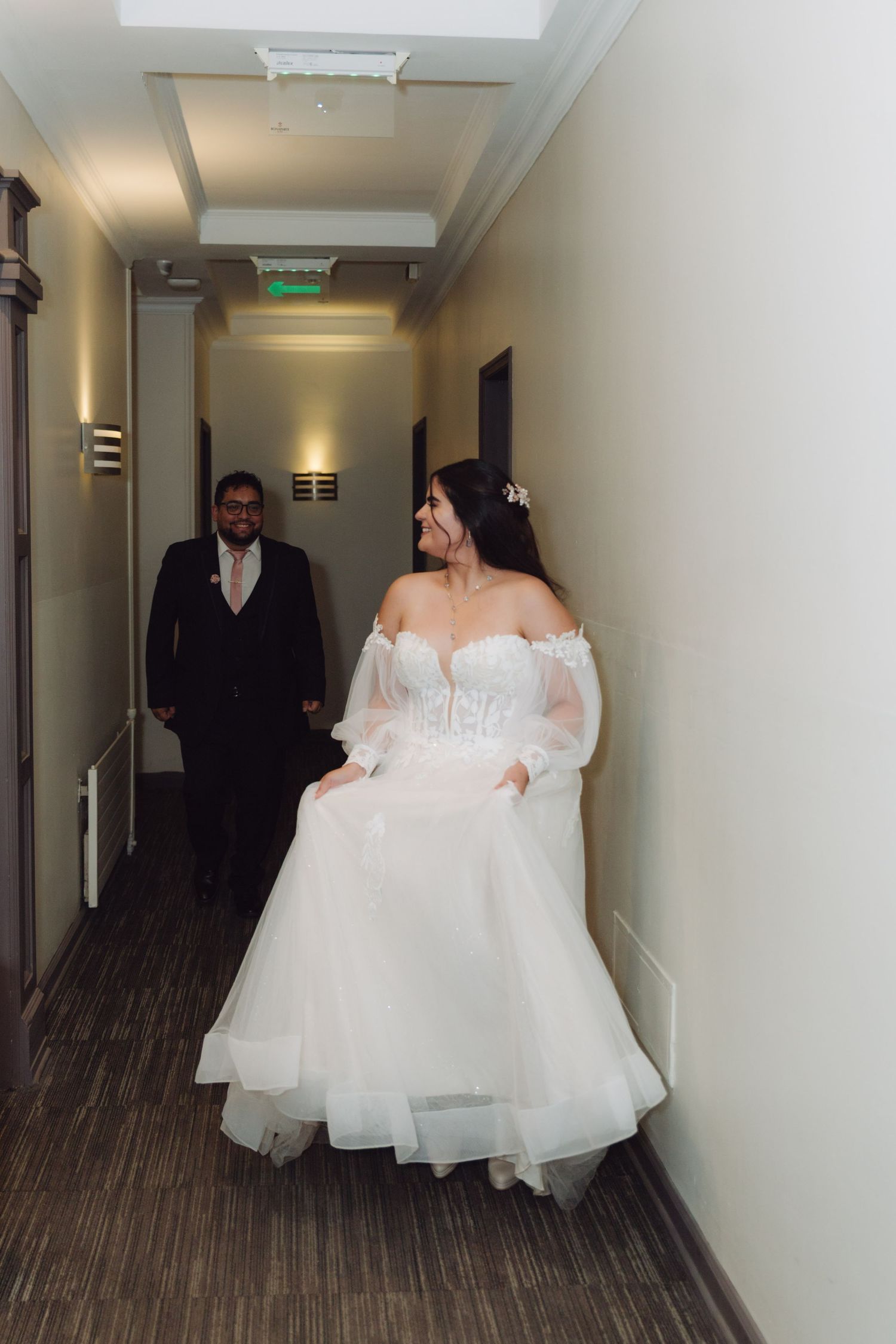 Radiant bride in white tulle gown walks hotel corridor with groom following behind.
