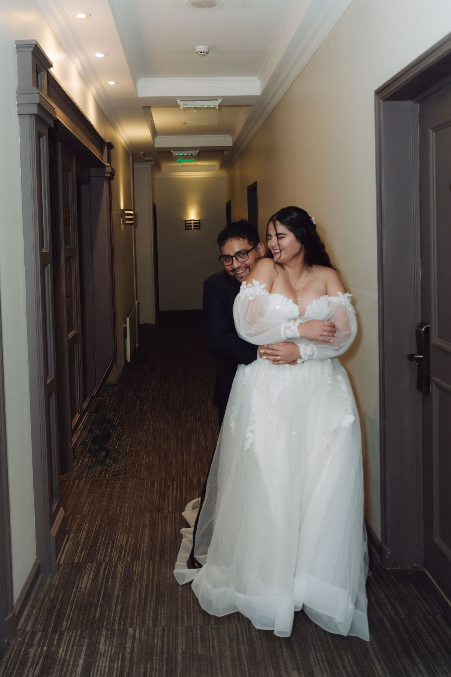 Newlywed couple shares a warm embrace and laugh together in hotel hallway.