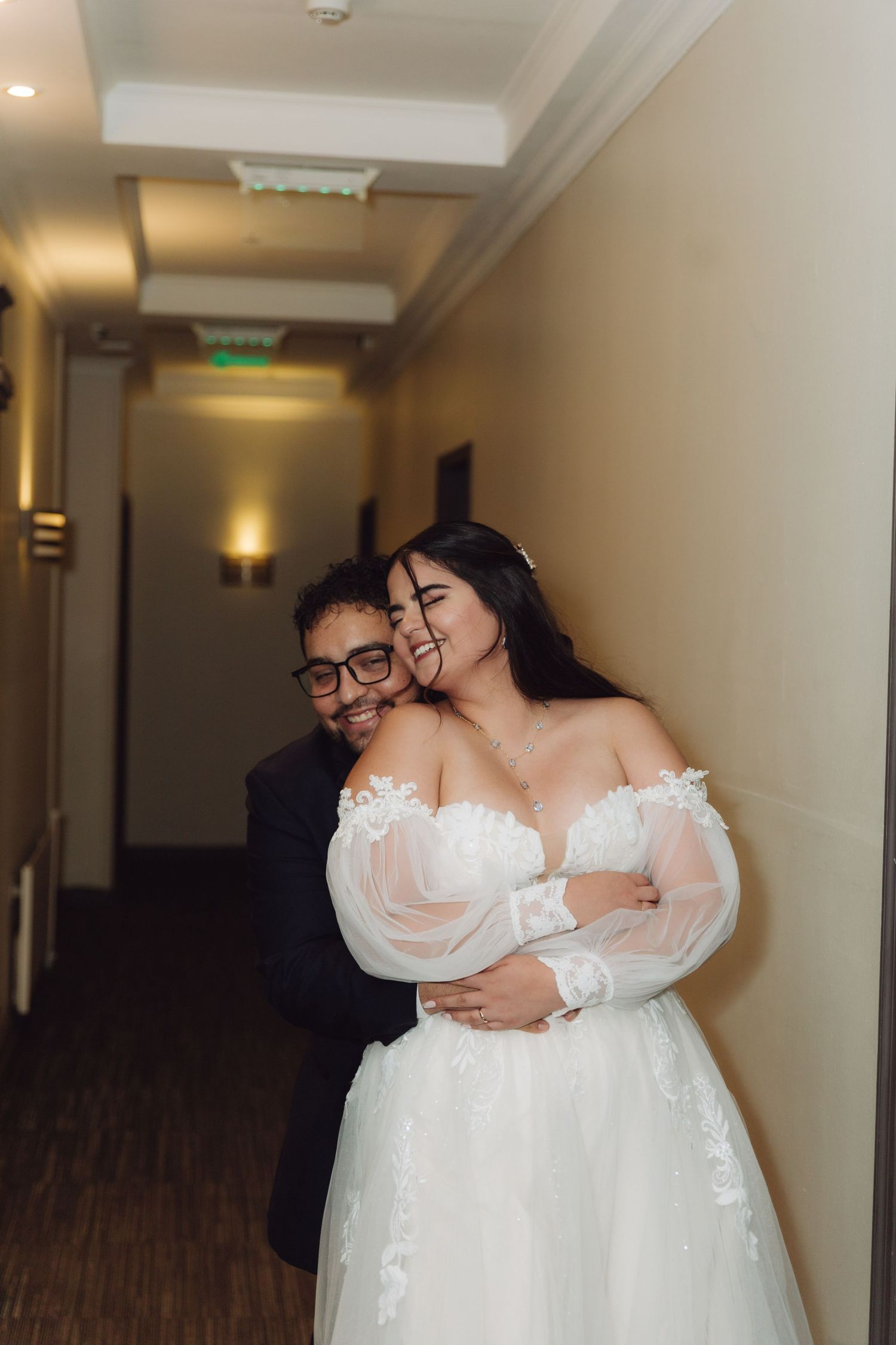 Happy bride and groom hug and smile together in warmly lit hotel corridor.