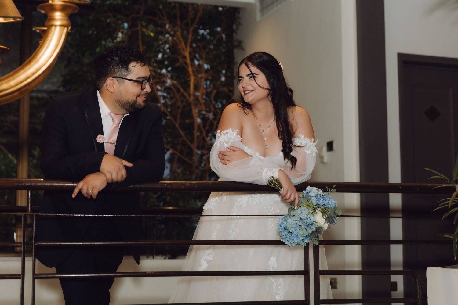 Elegant bride holding blue bouquet smiles at groom on decorative venue balcony.