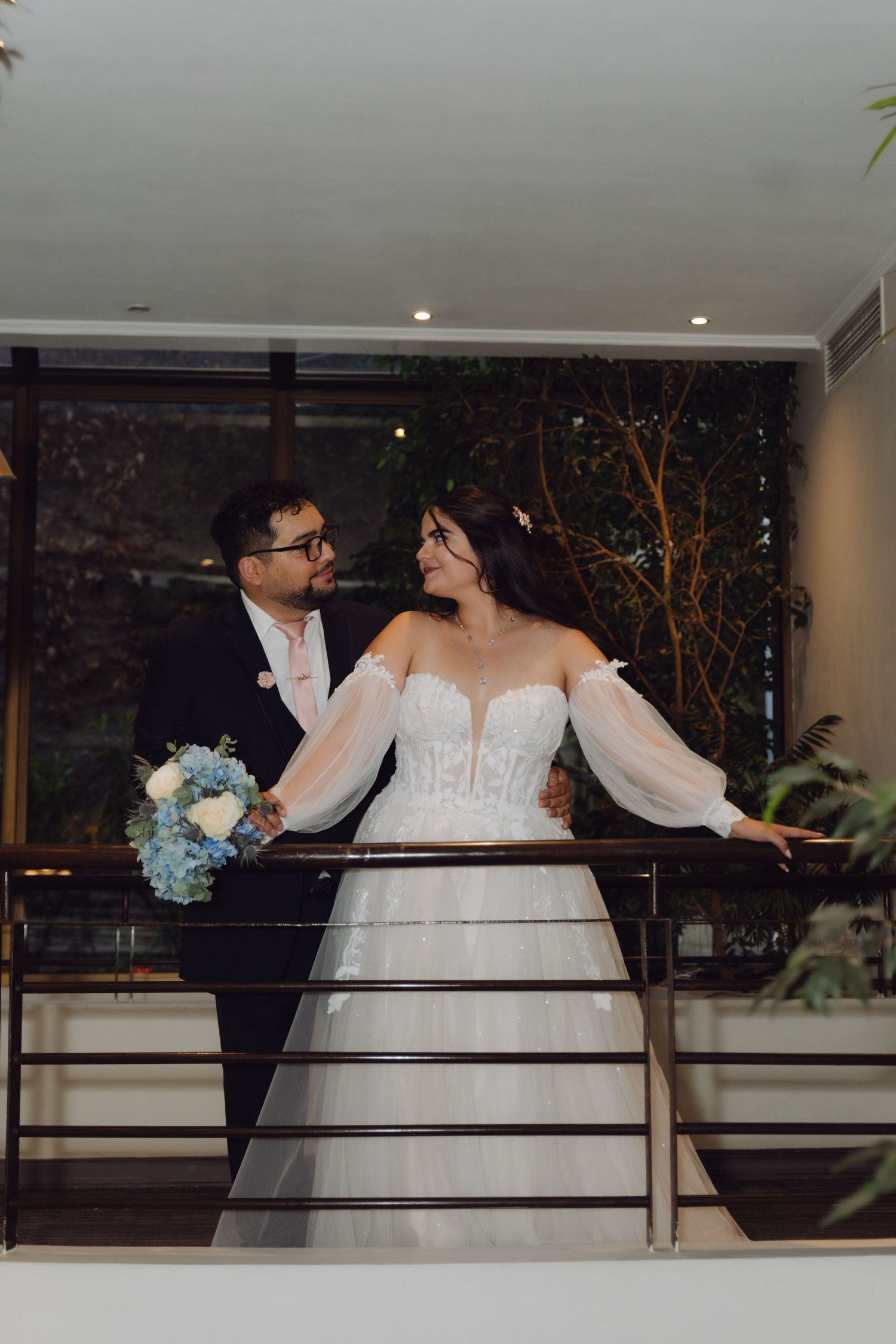Bride and groom share a romantic glance on balcony with lush greenery backdrop.