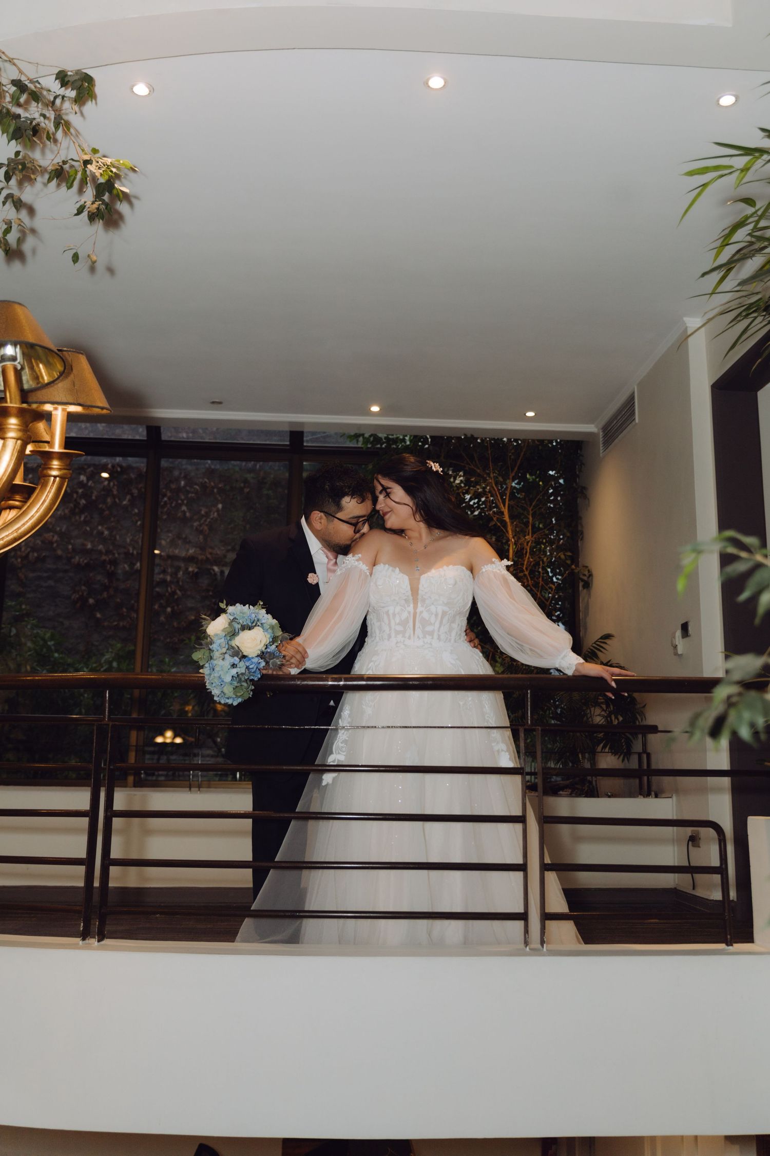 Newlyweds lean close together on balcony with blue bouquet at elegant wedding venue.