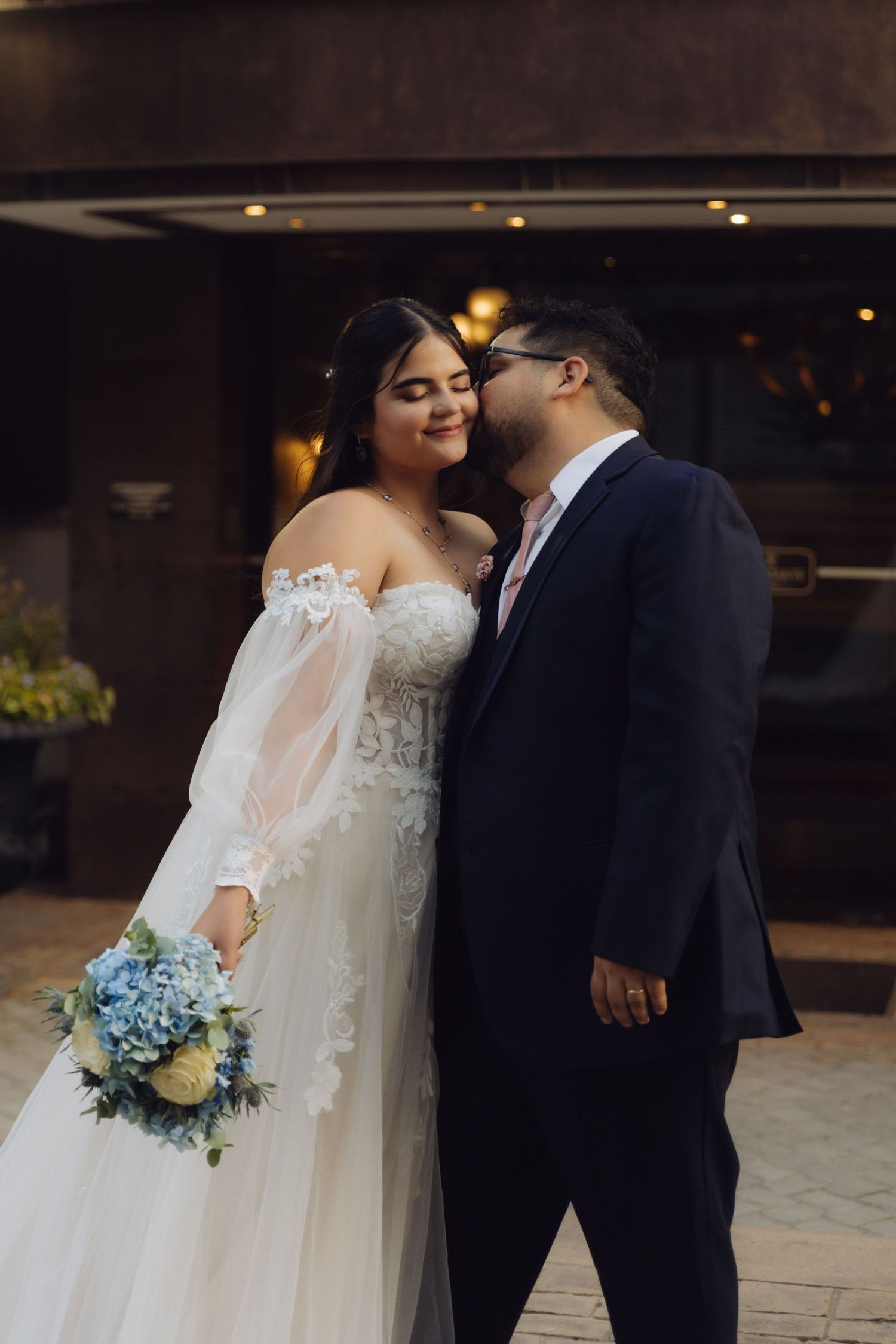 Bride and groom share an intimate moment, she holds blue hydrangea bouquet in white lace off-shoulder gown.