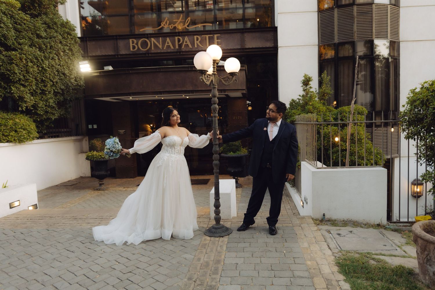 Newlyweds pose playfully at a lamppost outside the Bonaparte hotel at dusk.