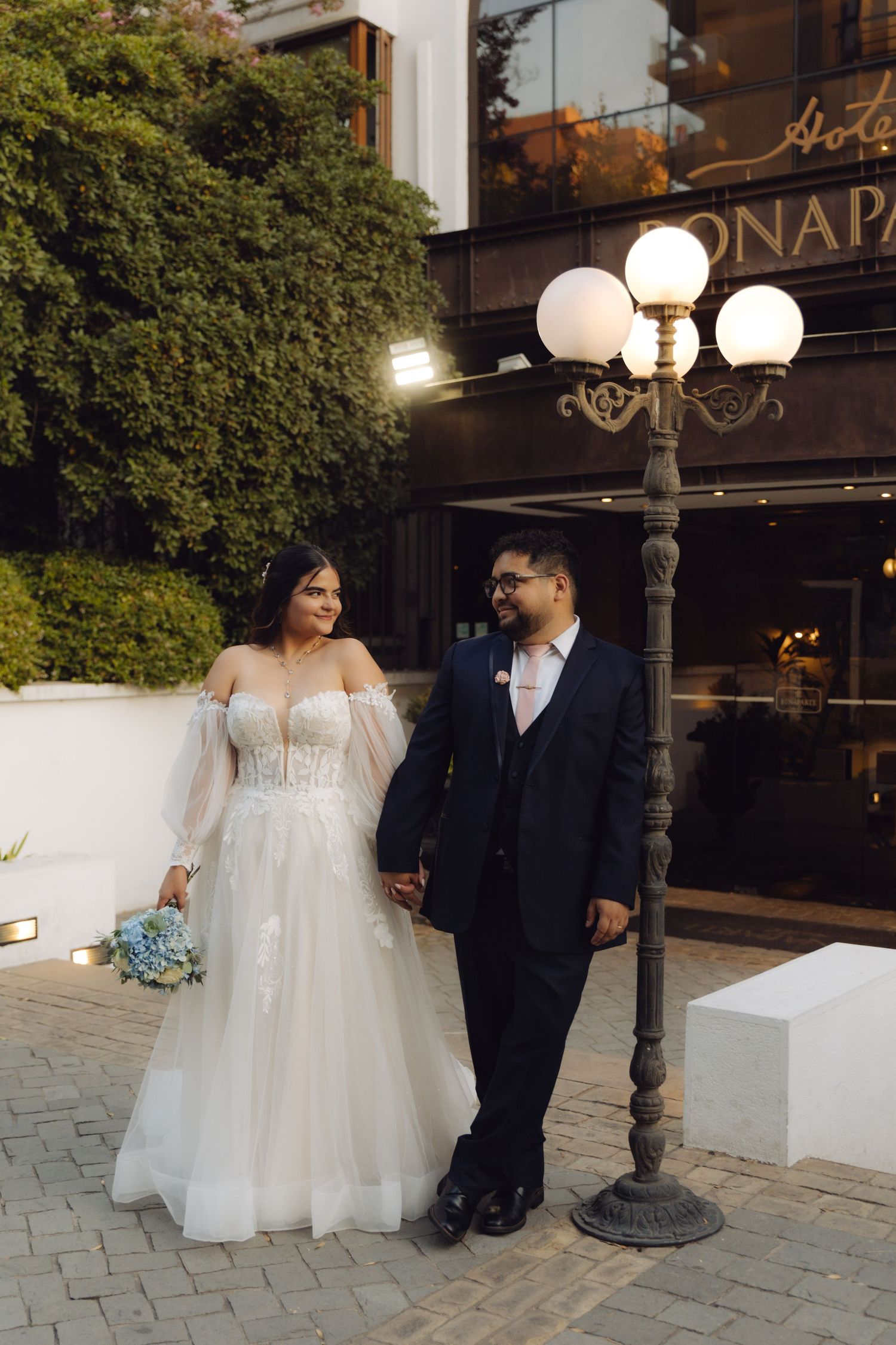 Bride and groom walk hand in hand outside Bonaparte hotel, bride carries blue hydrangea bouquet.