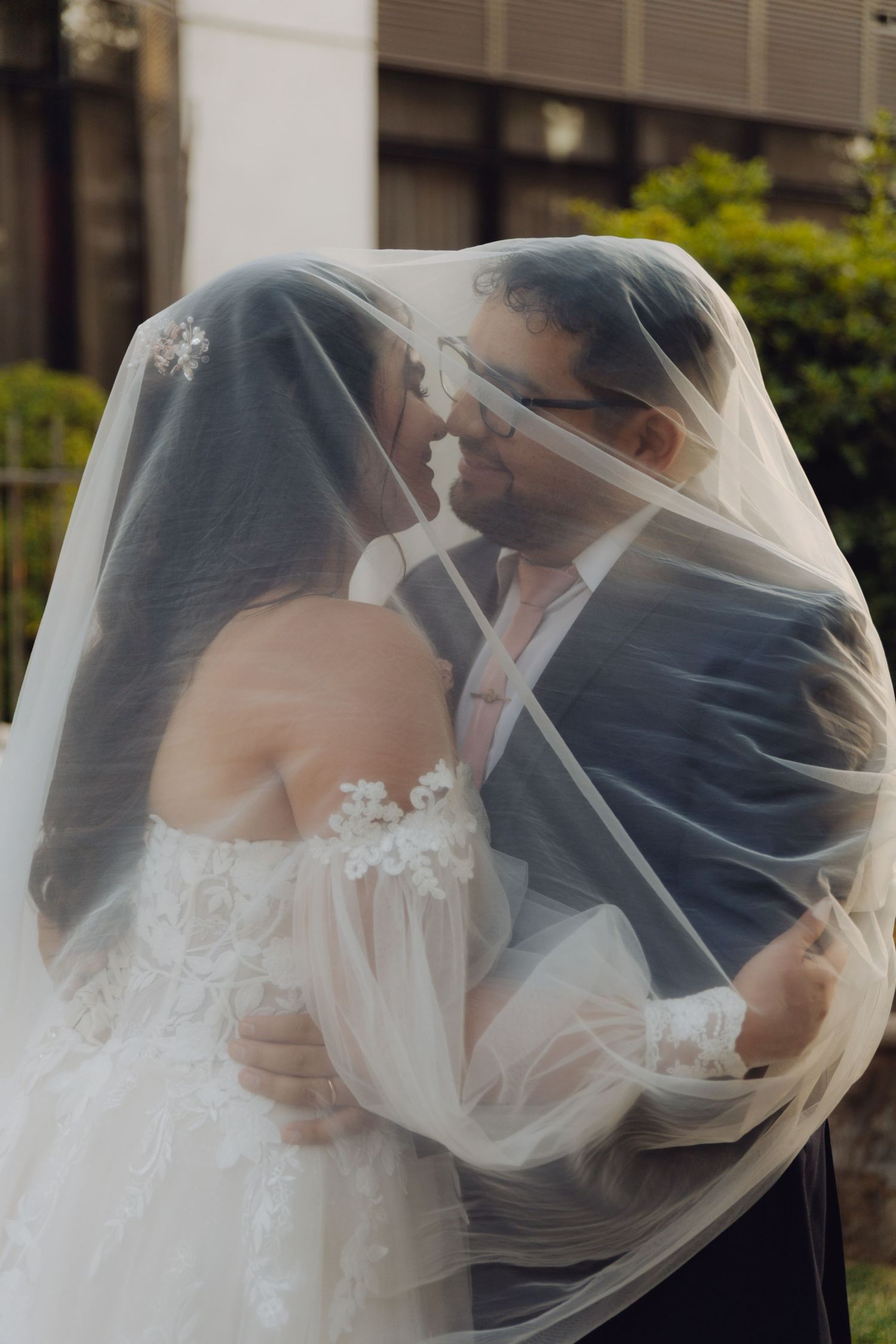 Romantic bride and groom share a kiss hidden beneath a flowing white wedding veil outdoors.