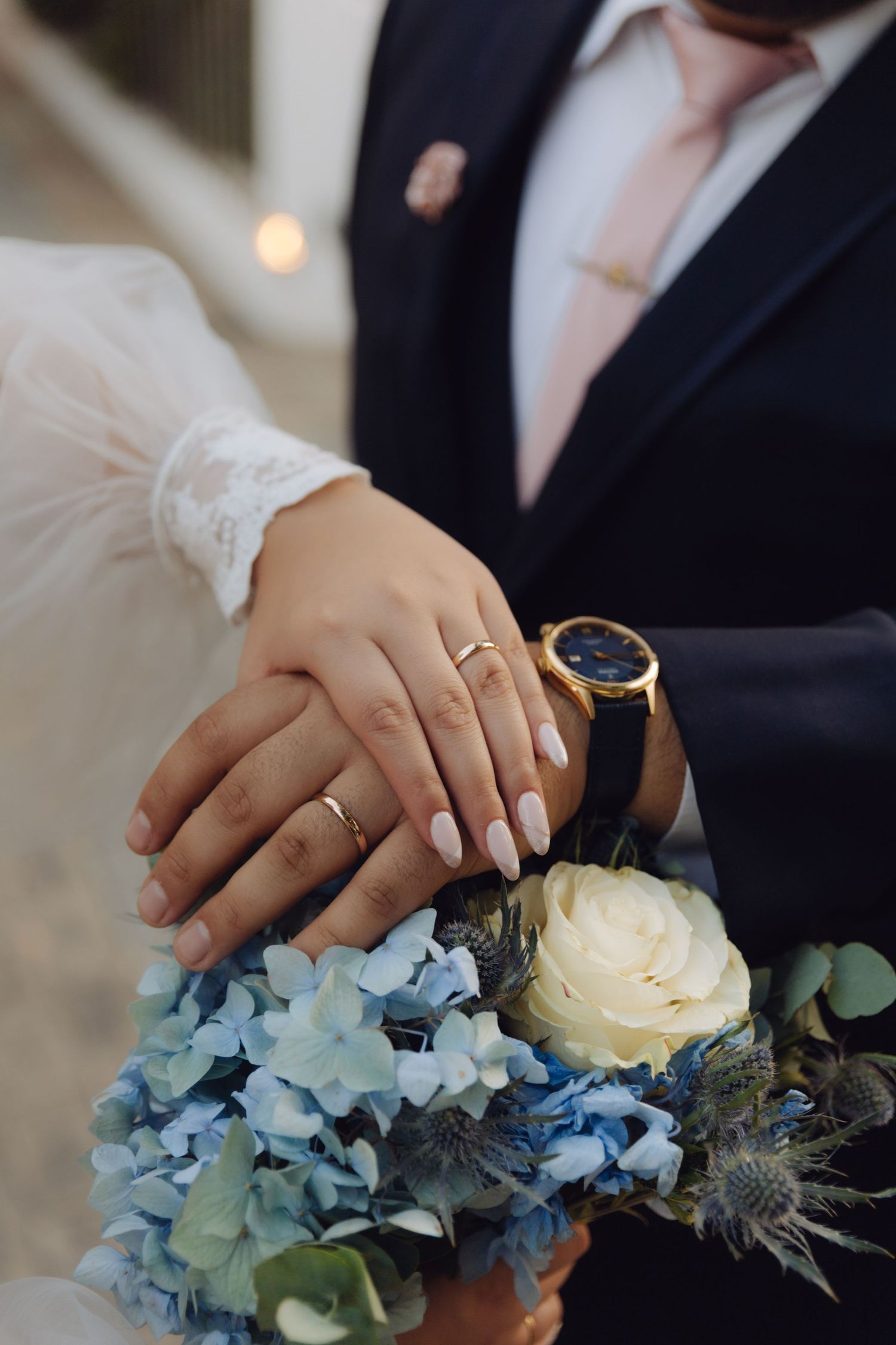 Close-up of newlyweds hands with wedding rings resting on blue hydrangea and white rose bouquet.