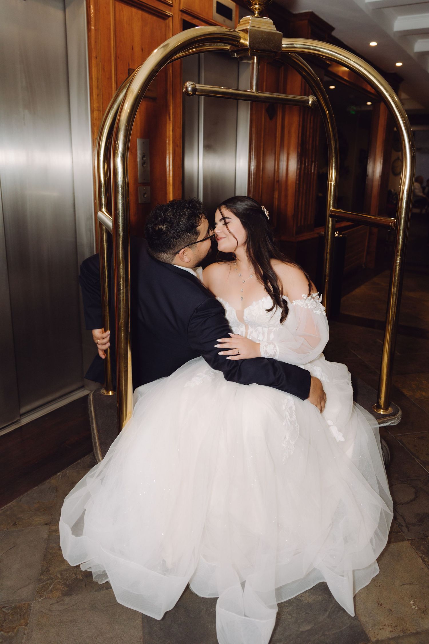 Bride and groom share a playful kiss on a gold hotel luggage cart in an elegant lobby.