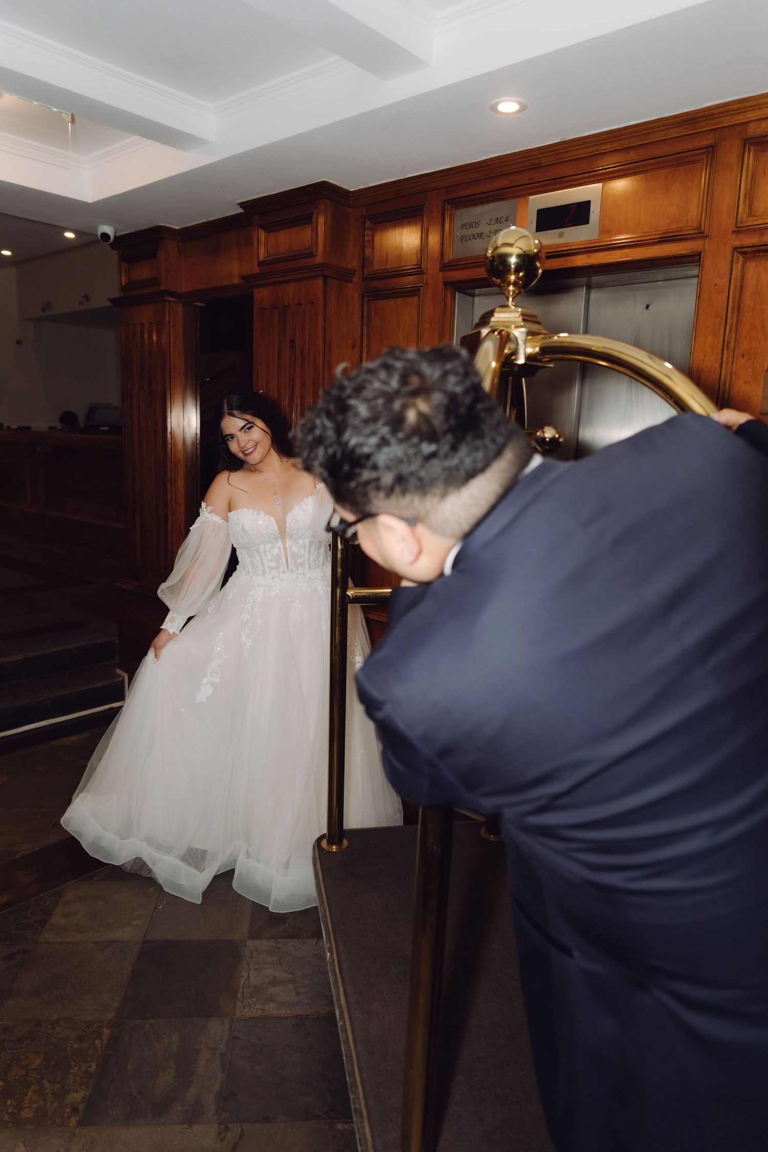 Groom reaches for bride near a gold luggage cart in a warmly lit hotel lobby interior.