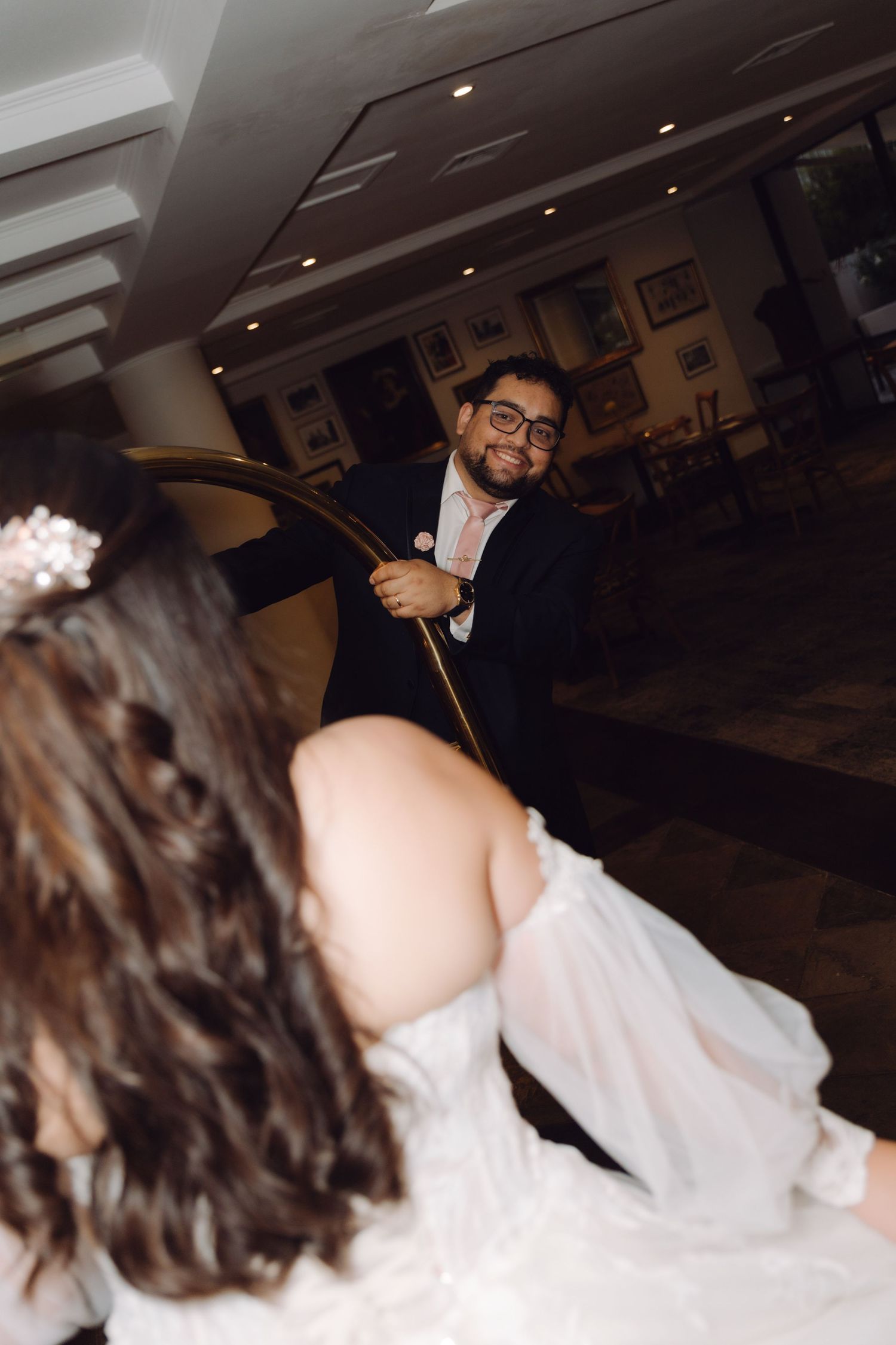 Smiling groom pulls bride on a gold luggage cart through a classic hotel lobby.