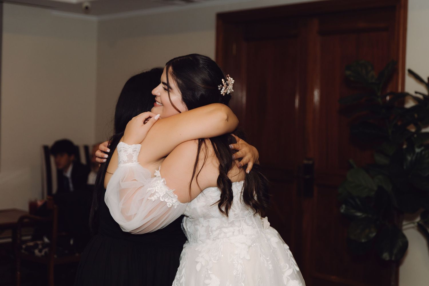 Bride in white lace gown shares a warm emotional hug with a female guest at the wedding.
