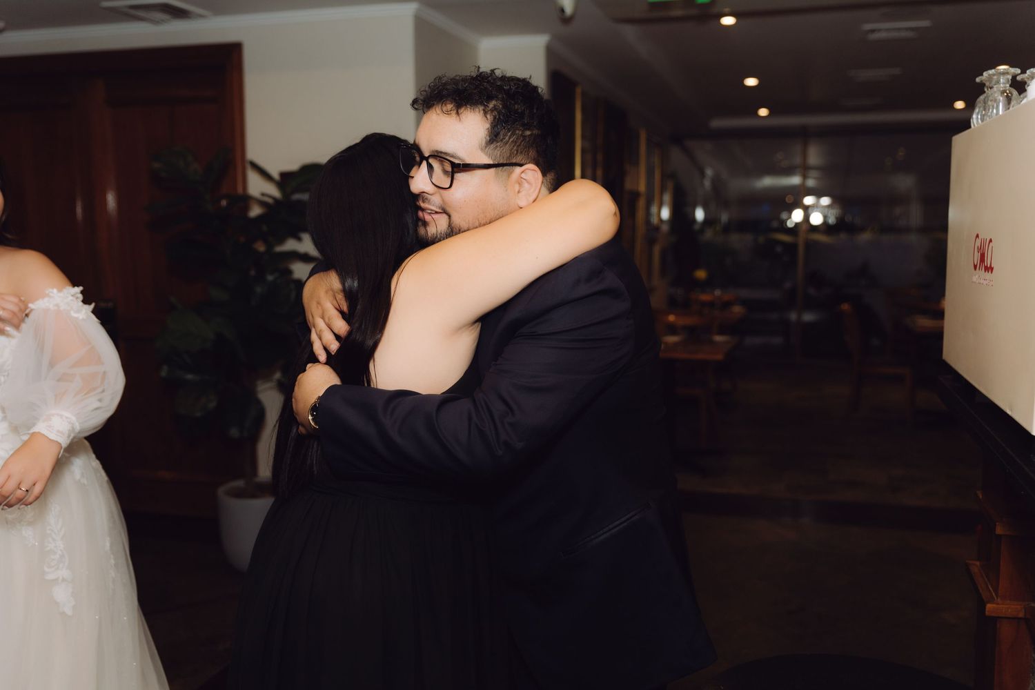Groom in navy suit shares a heartfelt embrace with a guest in a warmly lit wedding venue.