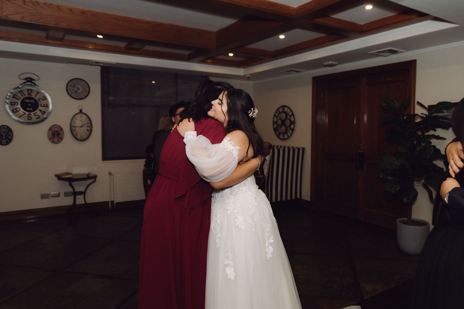 Bride in white gown embracing woman in burgundy dress at wedding reception indoors.