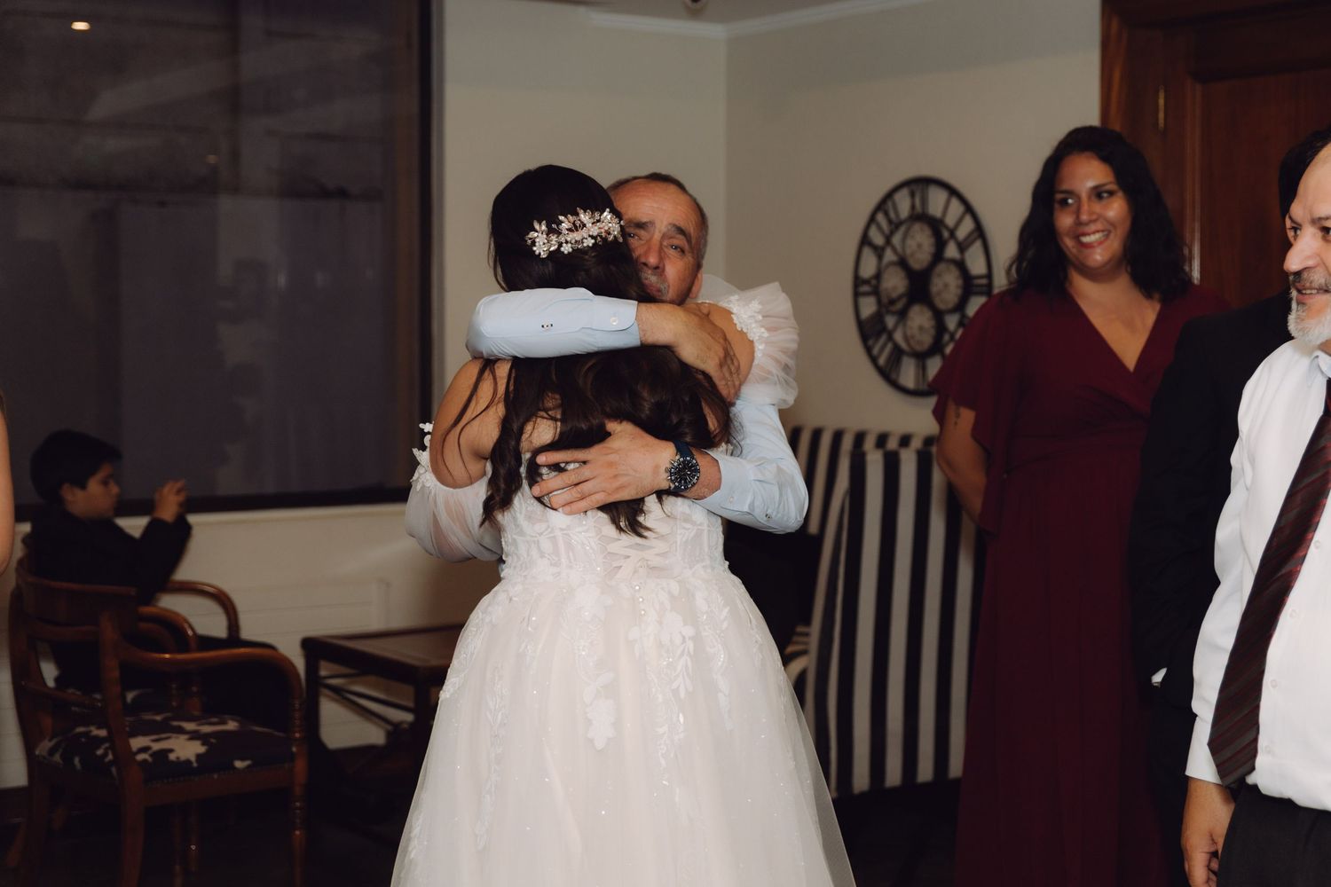 Bride in lace gown receiving warm hug from older man at wedding reception.