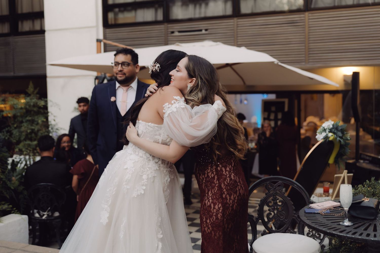 Bride in off-shoulder gown embracing woman in burgundy dress at outdoor wedding.