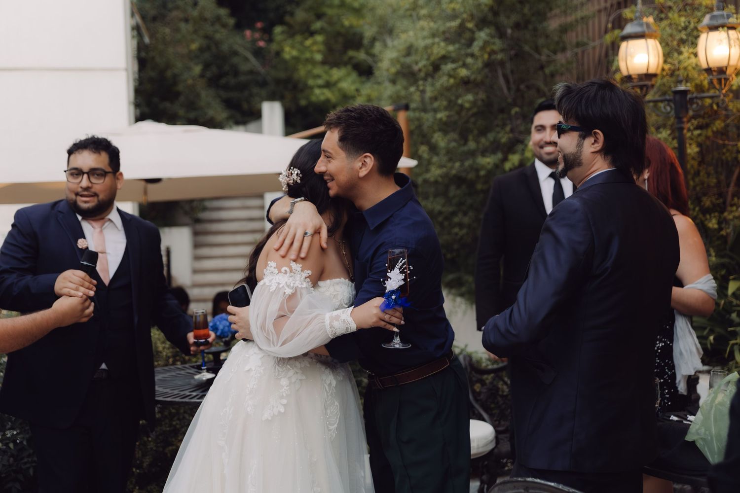 Bride in lace wedding dress embracing male guest at outdoor evening reception.