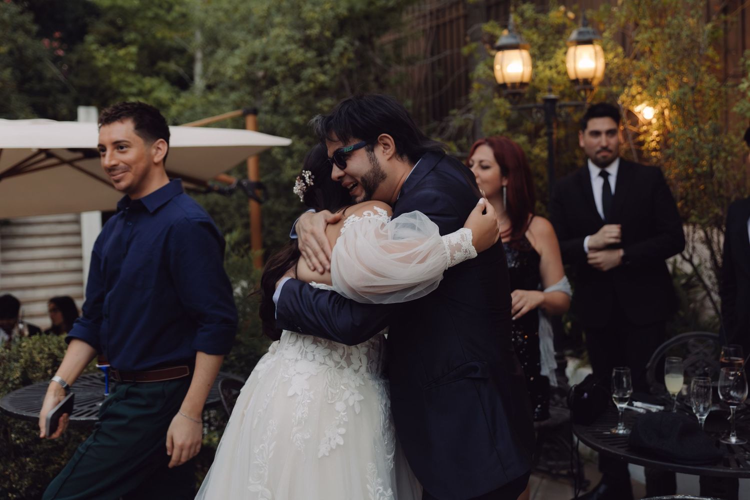 Bride in white gown sharing warm embrace with sunglasses-wearing guest at wedding.