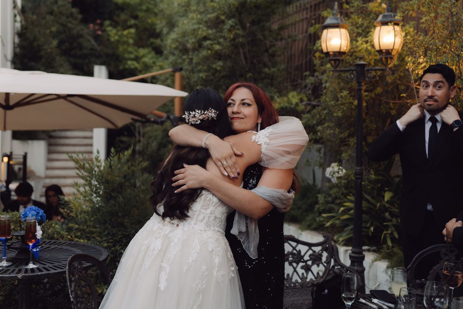 Bride with floral hair piece receiving hug from redhead woman at outdoor wedding.