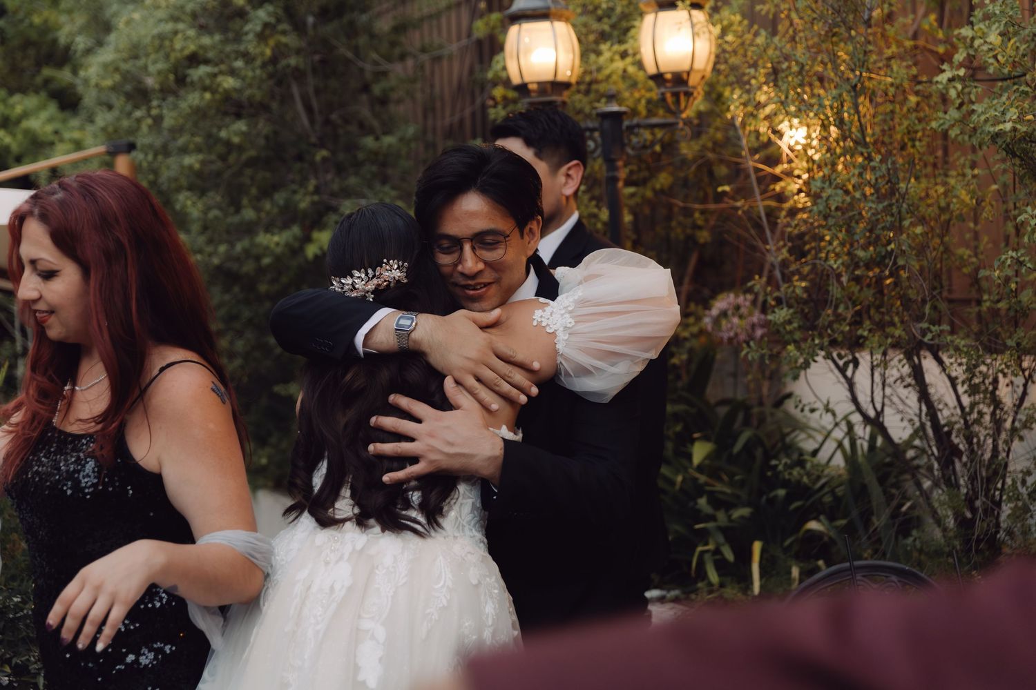 Bride in white lace gown embraced by couple at elegant outdoor wedding reception.