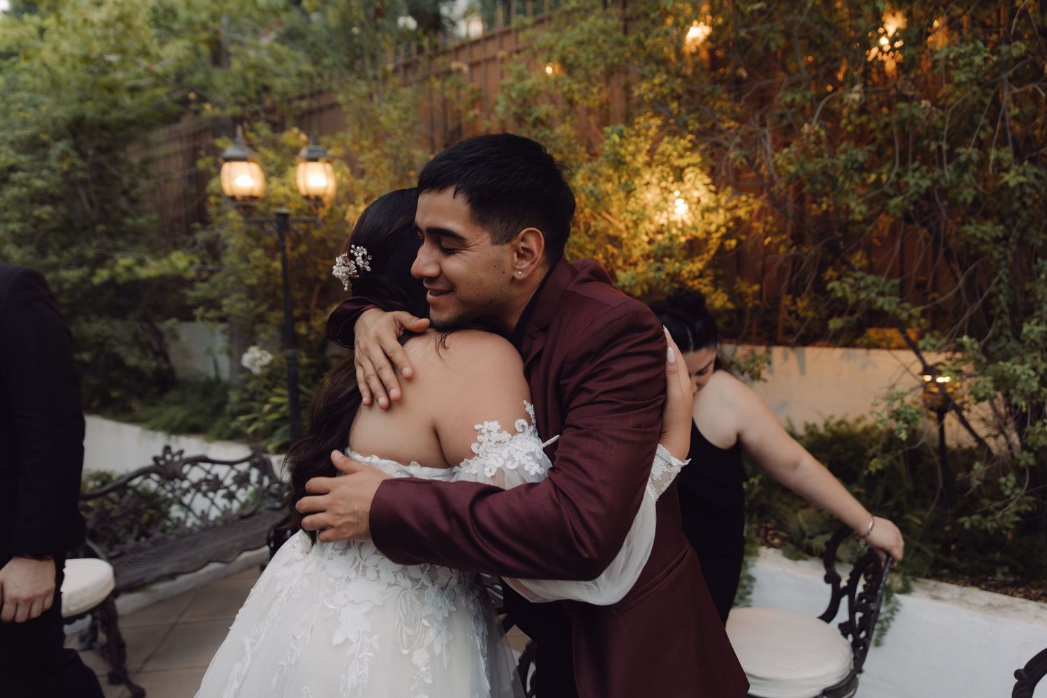 Bride and groom sharing a warm embrace at outdoor wedding reception surrounded by lush greenery and warm lights.