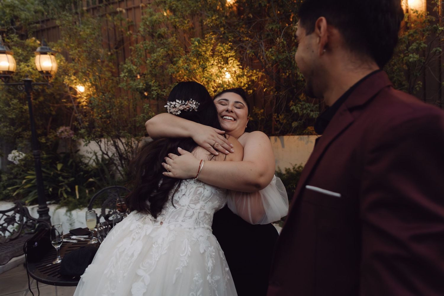 Joyful bride hugging wedding guest while groom in burgundy suit watches at elegant outdoor reception.