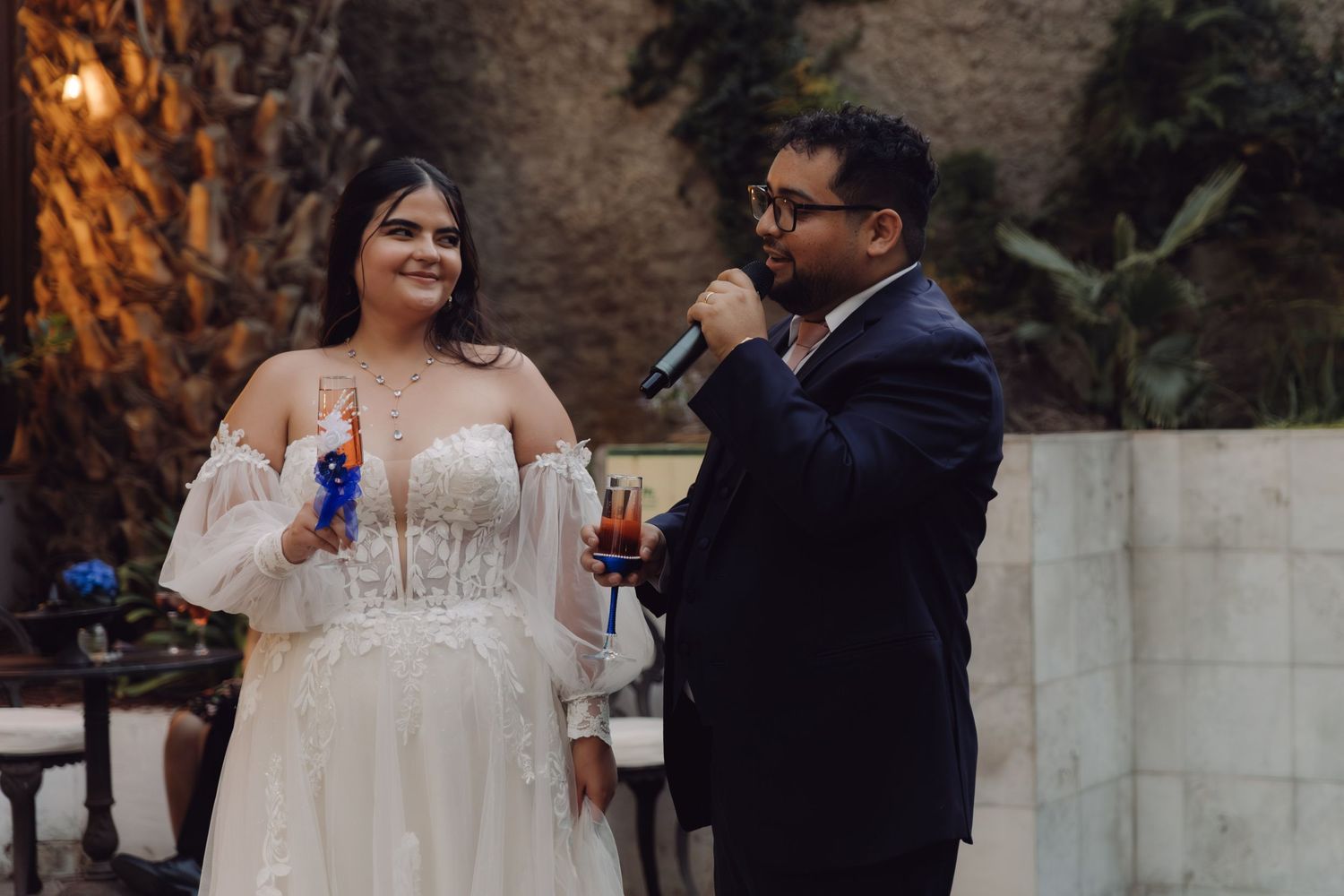 Bride in off-shoulder lace gown and groom in navy suit speaking into microphone during wedding toast.