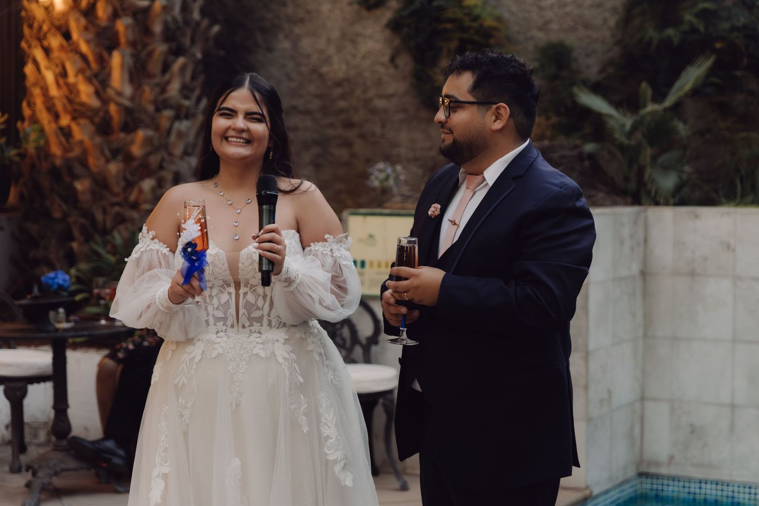 Smiling bride and groom holding champagne glasses during wedding reception toast at outdoor venue.